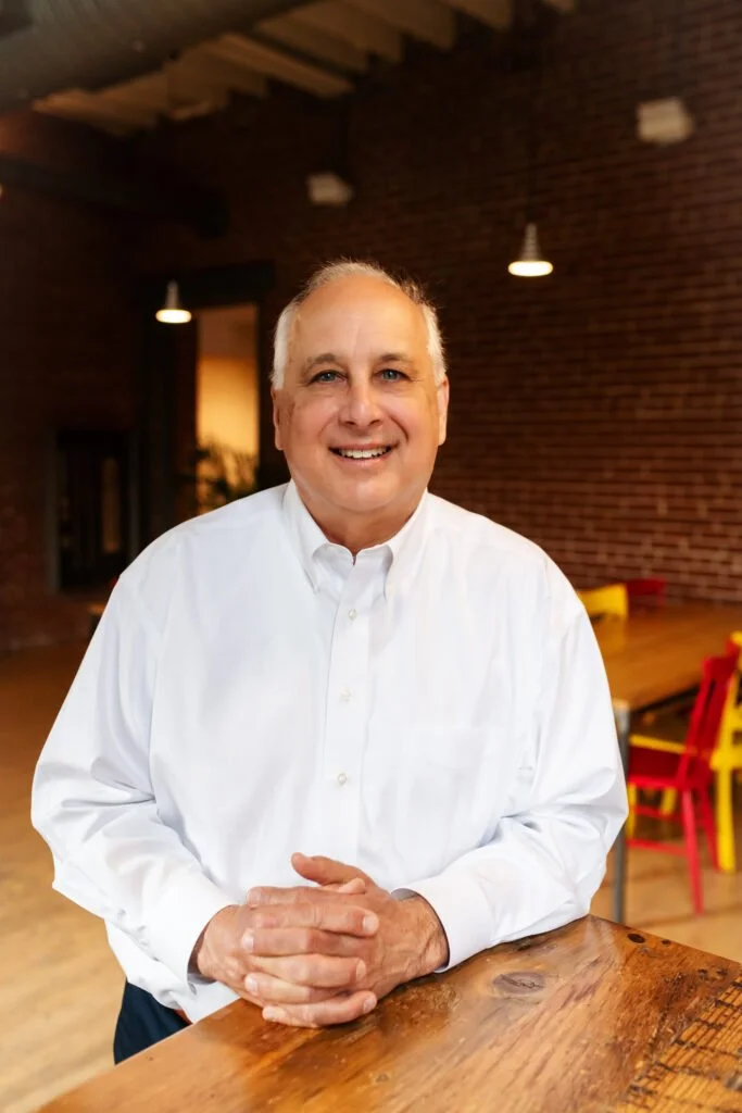 A smiling middle-aged man with white hair, wearing a white button-up shirt, standing in a warmly lit room with brick walls and colorful chairs in the background.
