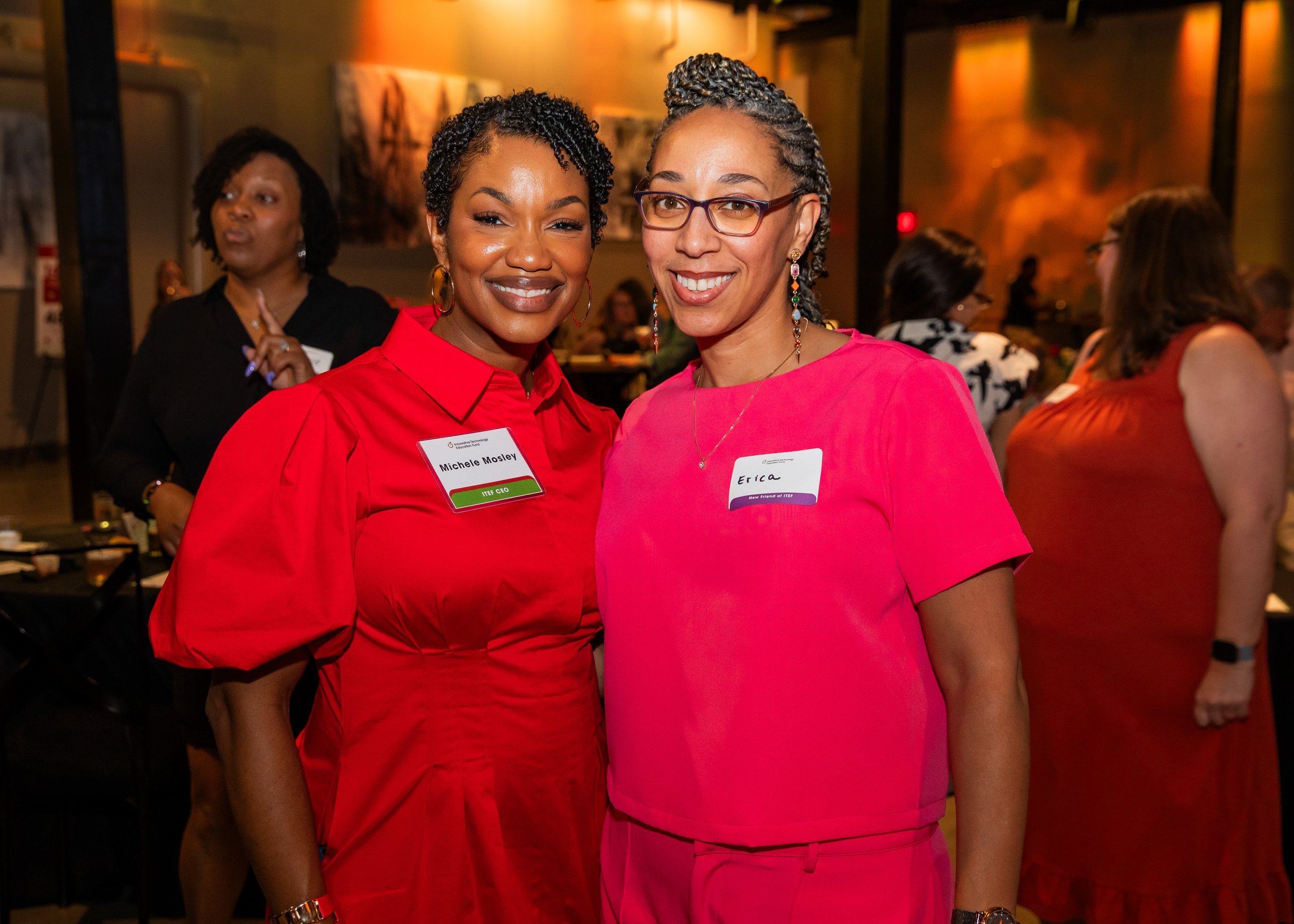 Two women wearing bright pink tops smiling at a social event, with other people in the background.