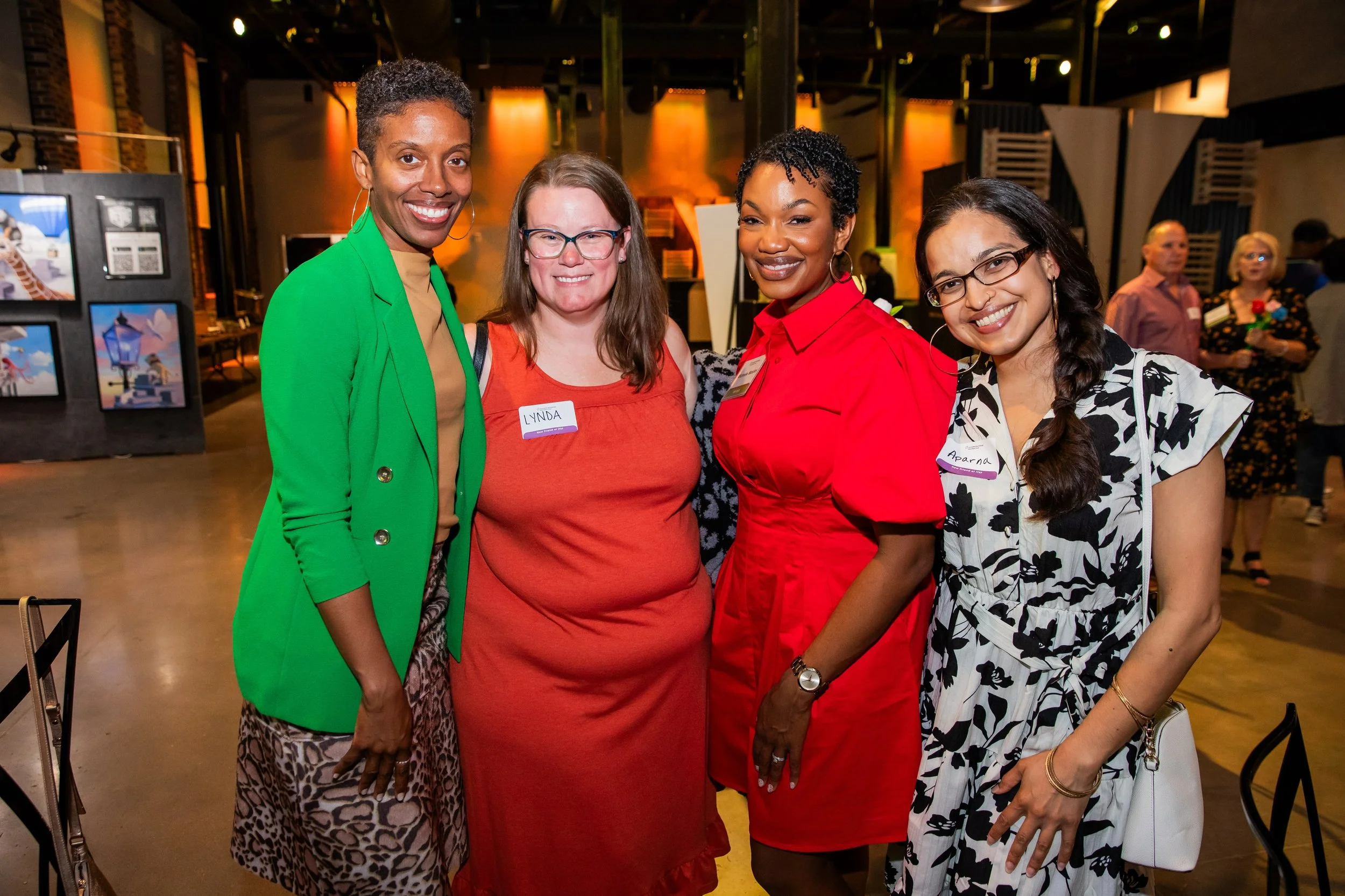 Four women smiling at an indoor event, three wearing colorful dresses and one in a black and white floral dress, in front of a group of people and artwork.