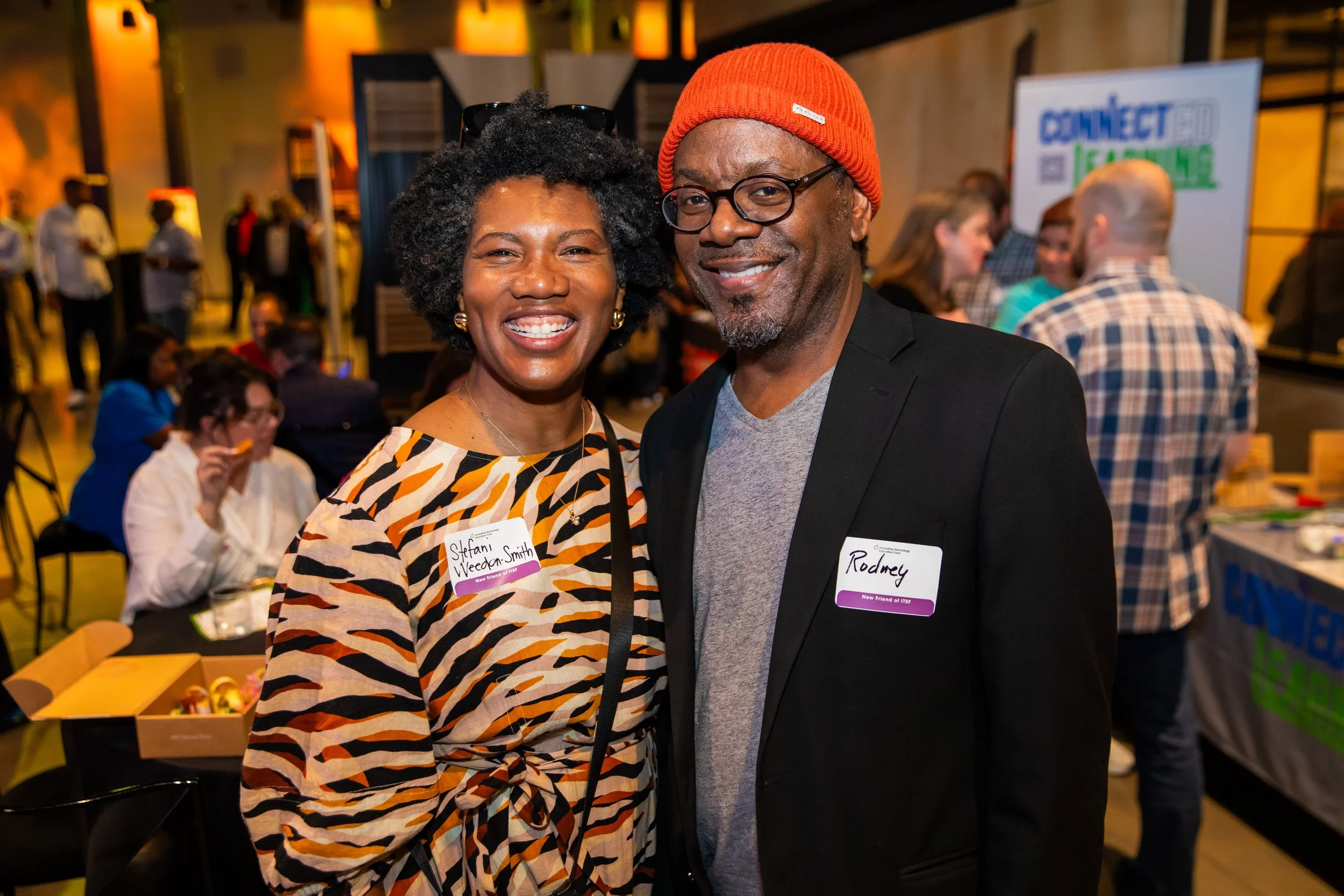 A smiling Black woman with curly hair wearing a colorful patterned dress and glasses on her head, standing next to a smiling Black man wearing a red beanie, glasses, a black blazer, and a gray shirt, at an indoor event with other people in the backgr
