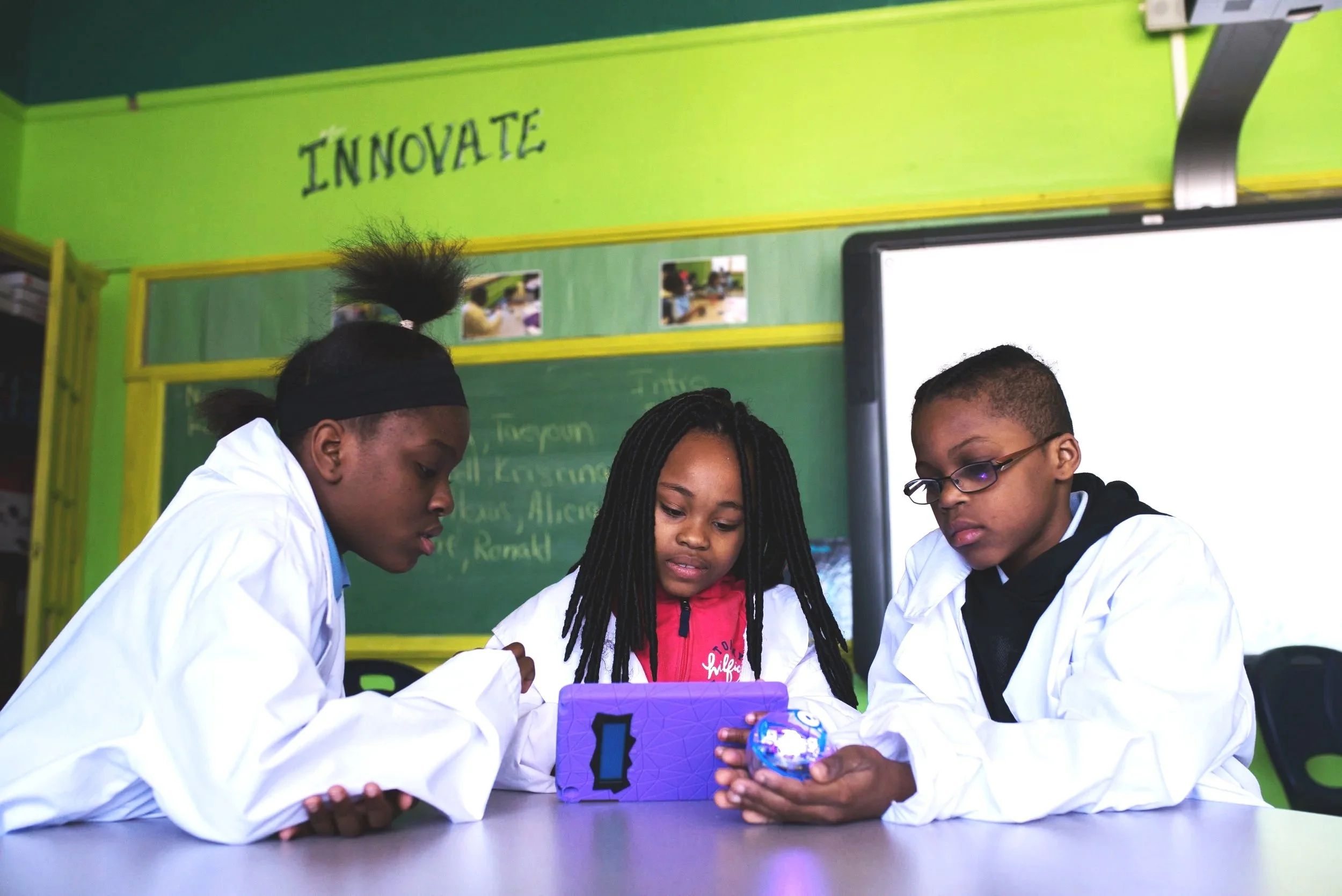 Three children in white coats sitting at a table together, focused on a purple tablet. A classroom setting with a green wall and the word 'INNOVATE' written on it is in the background.