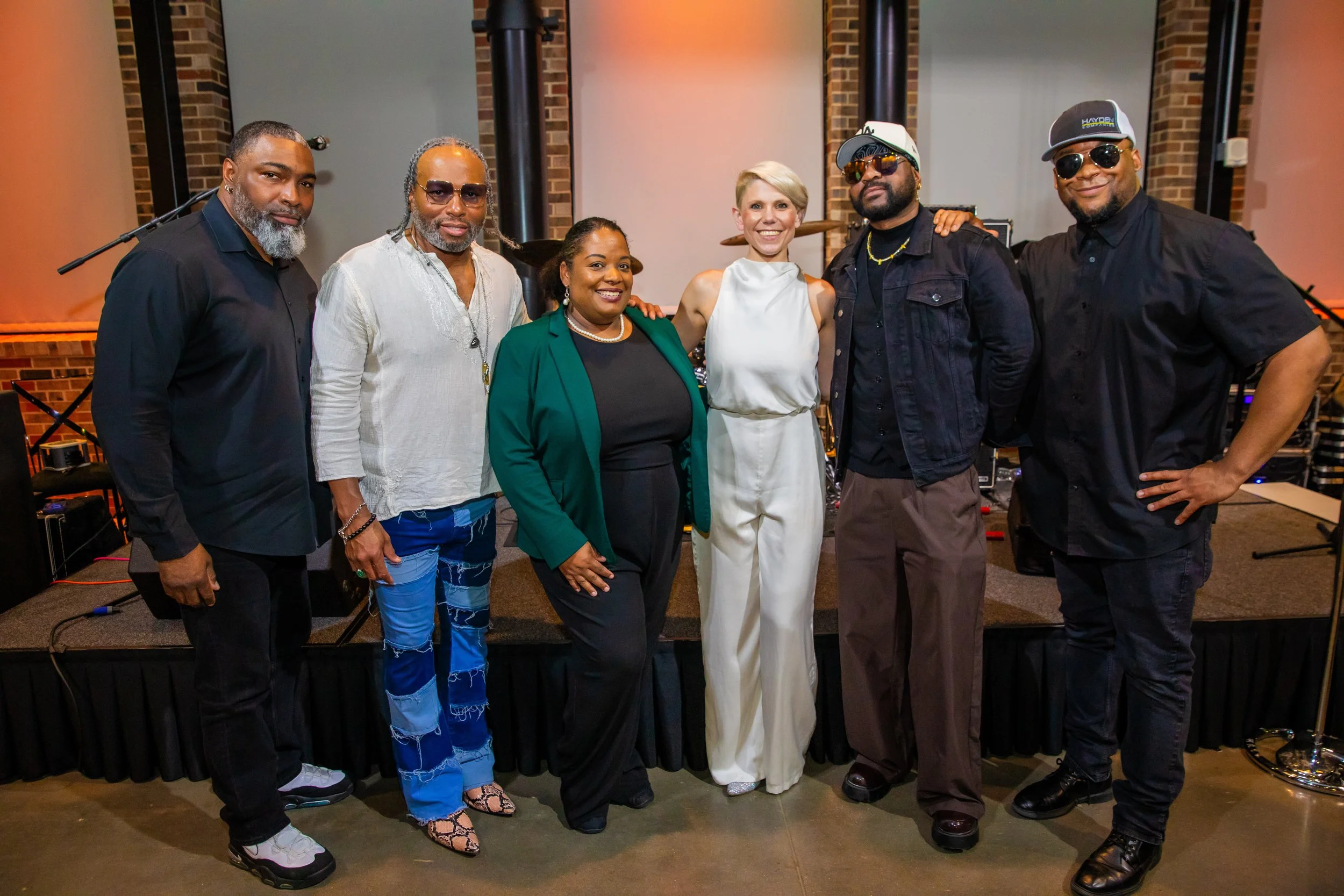 Group of six diverse adults standing together indoors, posing for a photo with musical equipment in the background.