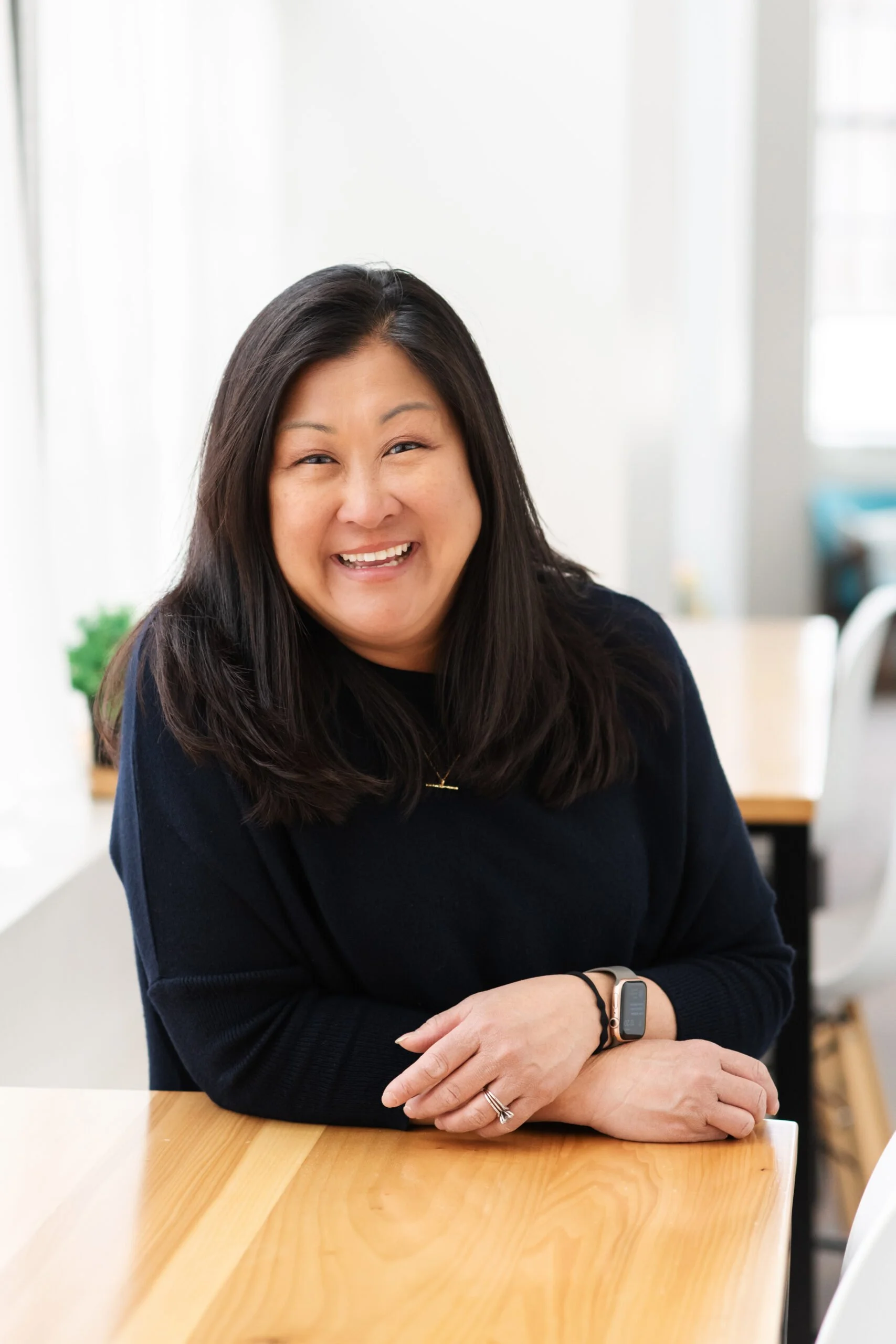 A woman with long dark hair smiling, wearing a black top and smartwatch, sitting at a wooden table in a bright, modern office environment.
