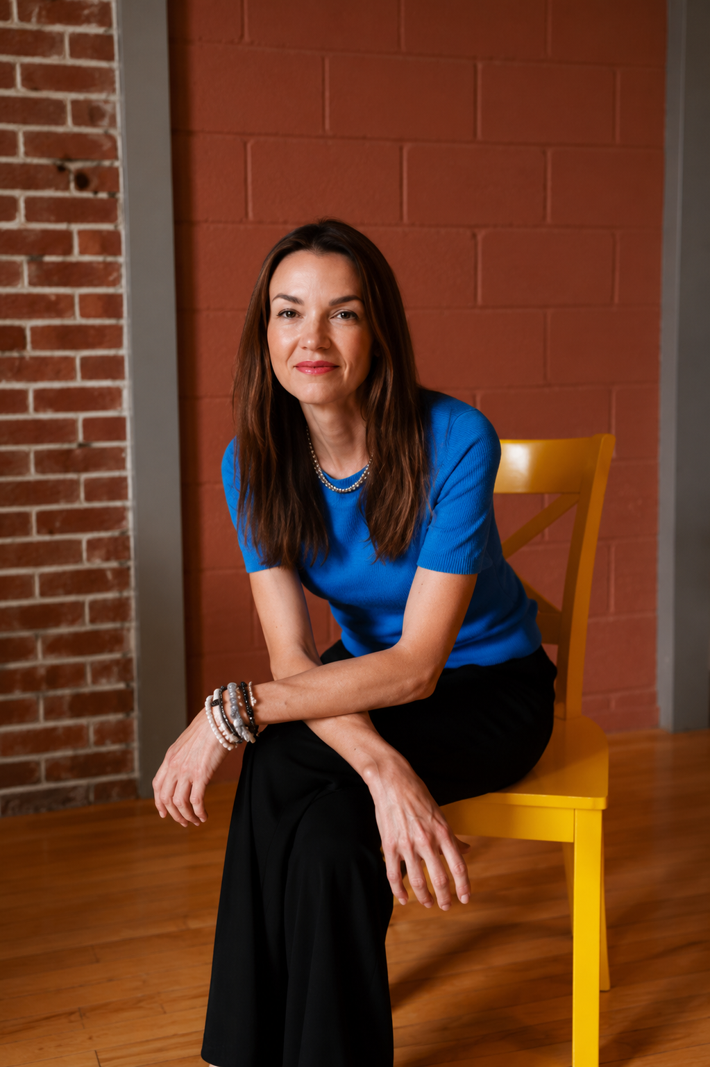 A woman with brown hair wearing a blue top, black pants, and multiple bracelets and a pearl necklace, sitting on a yellow chair in front of a brick and painted wall.