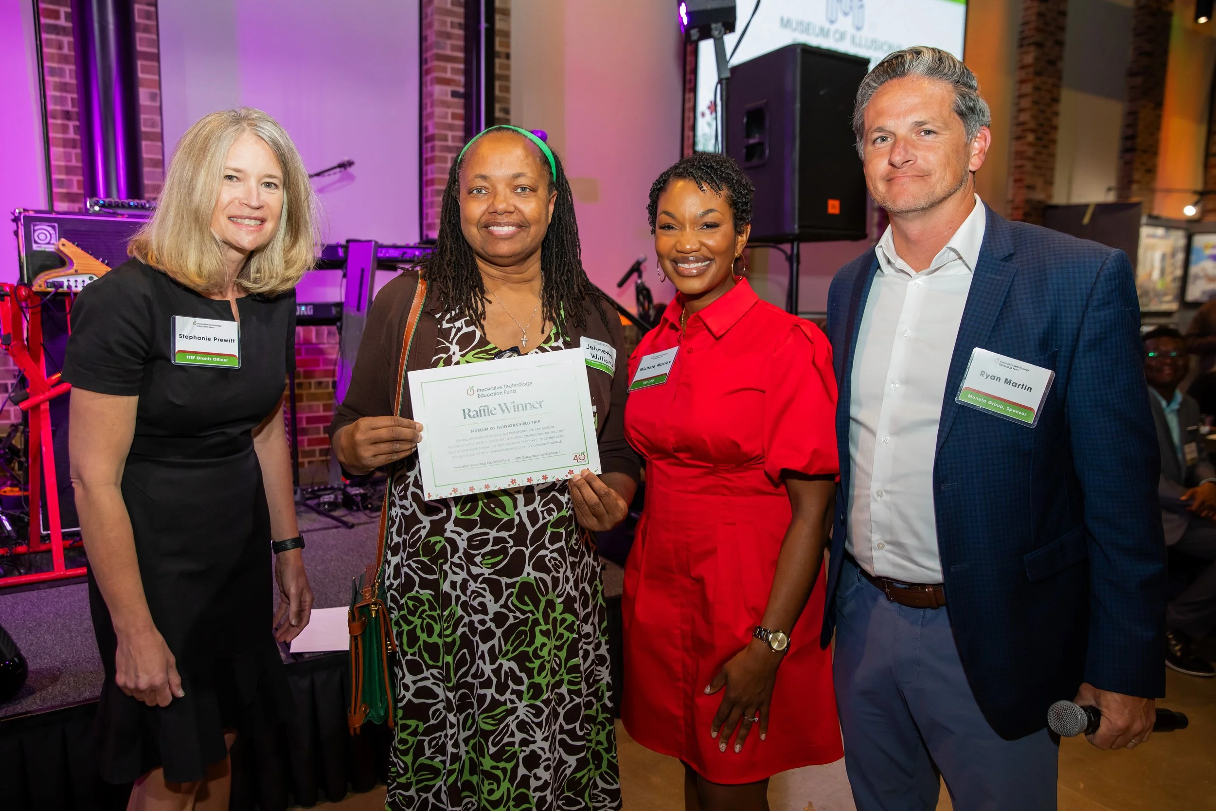 Five people standing together at an indoor event, smiling at the camera. The woman in the center holds a certificate. They wear business casual attire, with name tags visible. The background has purple and orange lighting, musical instruments, and a 