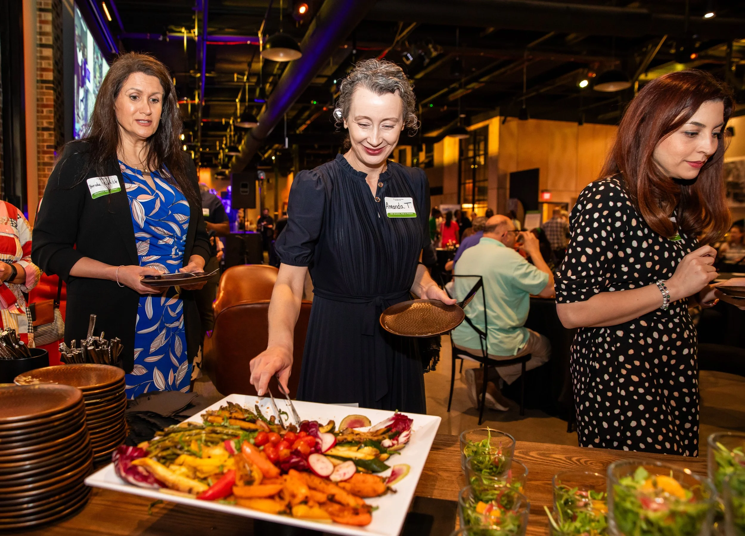 Three women standing at a buffet table with plates of food and salad bowls in a warmly lit restaurant or event space, with other guests in the background.