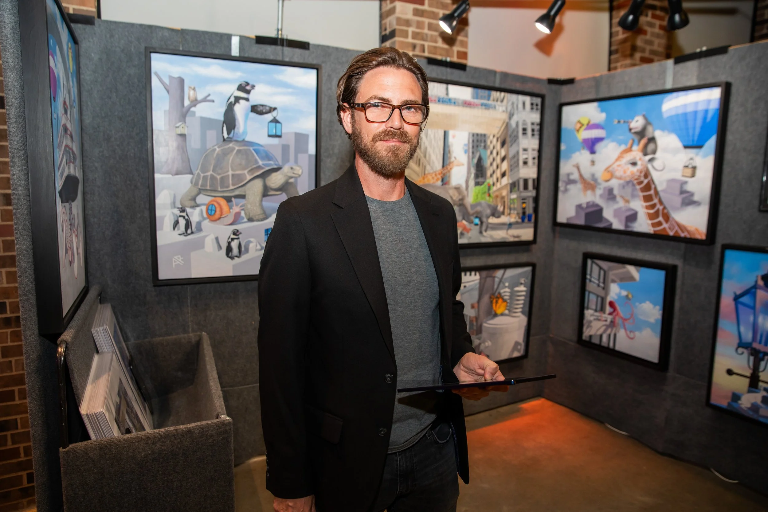 A man with glasses and a beard in a black blazer at an art gallery, surrounded by colorful animated-style illustrations of animals and cityscapes displayed on screens.