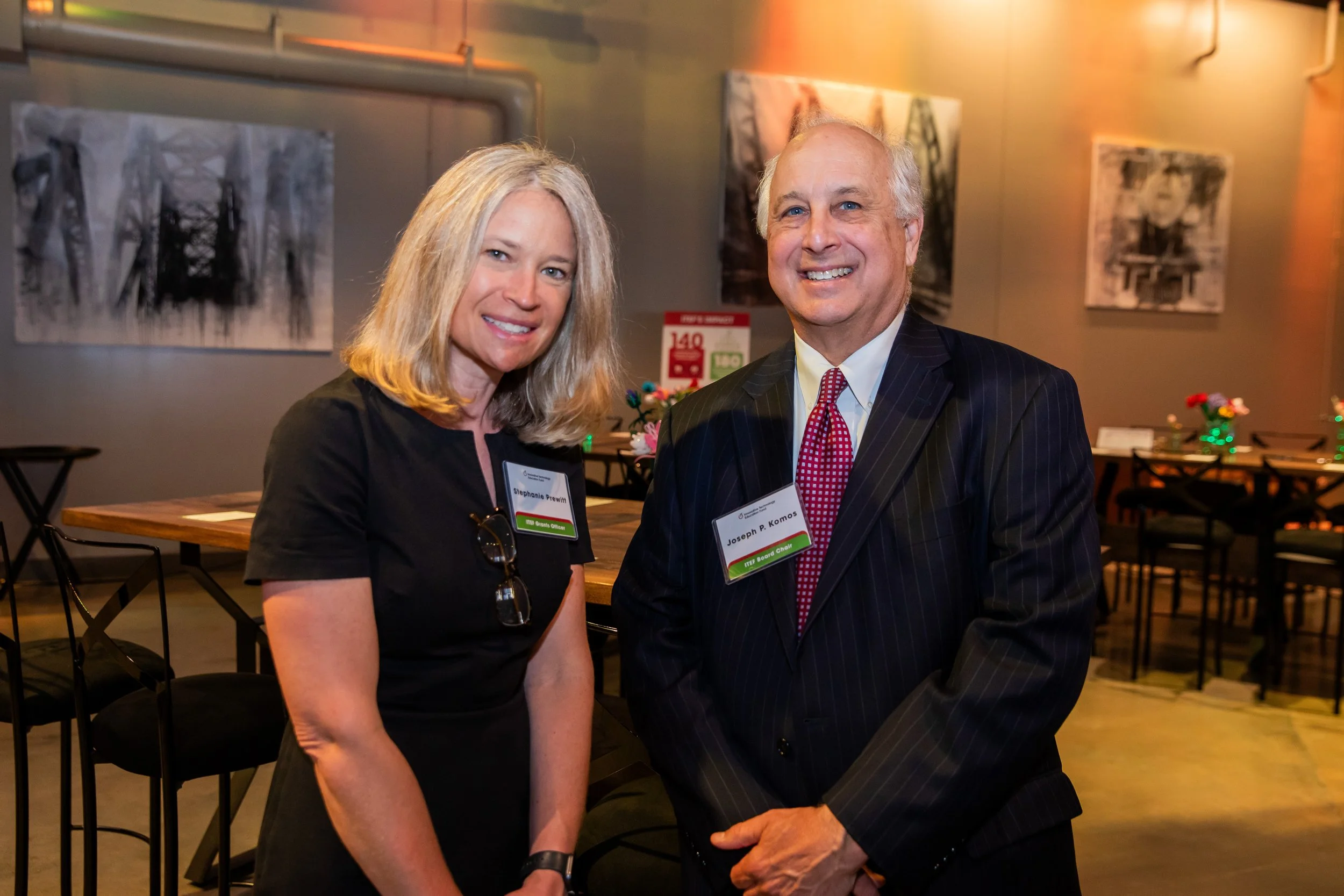 A man and a woman standing side by side in an art gallery, smiling at the camera. The woman has blonde hair and is wearing a black top, and the man has gray hair and is dressed in a suit with a white shirt and red tie. They are wearing name tags, and