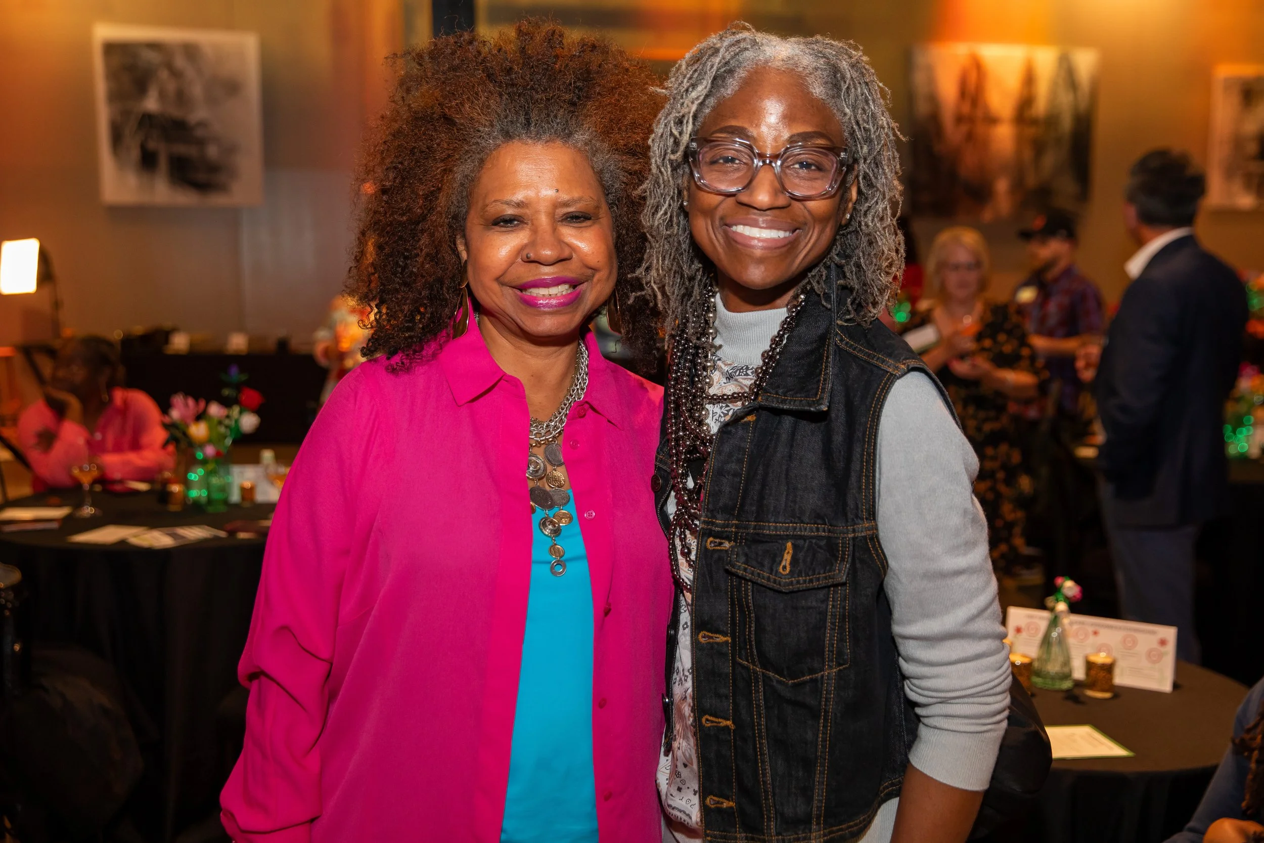 Two women smiling at a social gathering, standing closely together. The woman on the left has curly gray hair, is wearing a bright pink blazer over a blue top, and jewelry. The woman on the right has gray dreadlocks, glasses, a denim vest over a patt