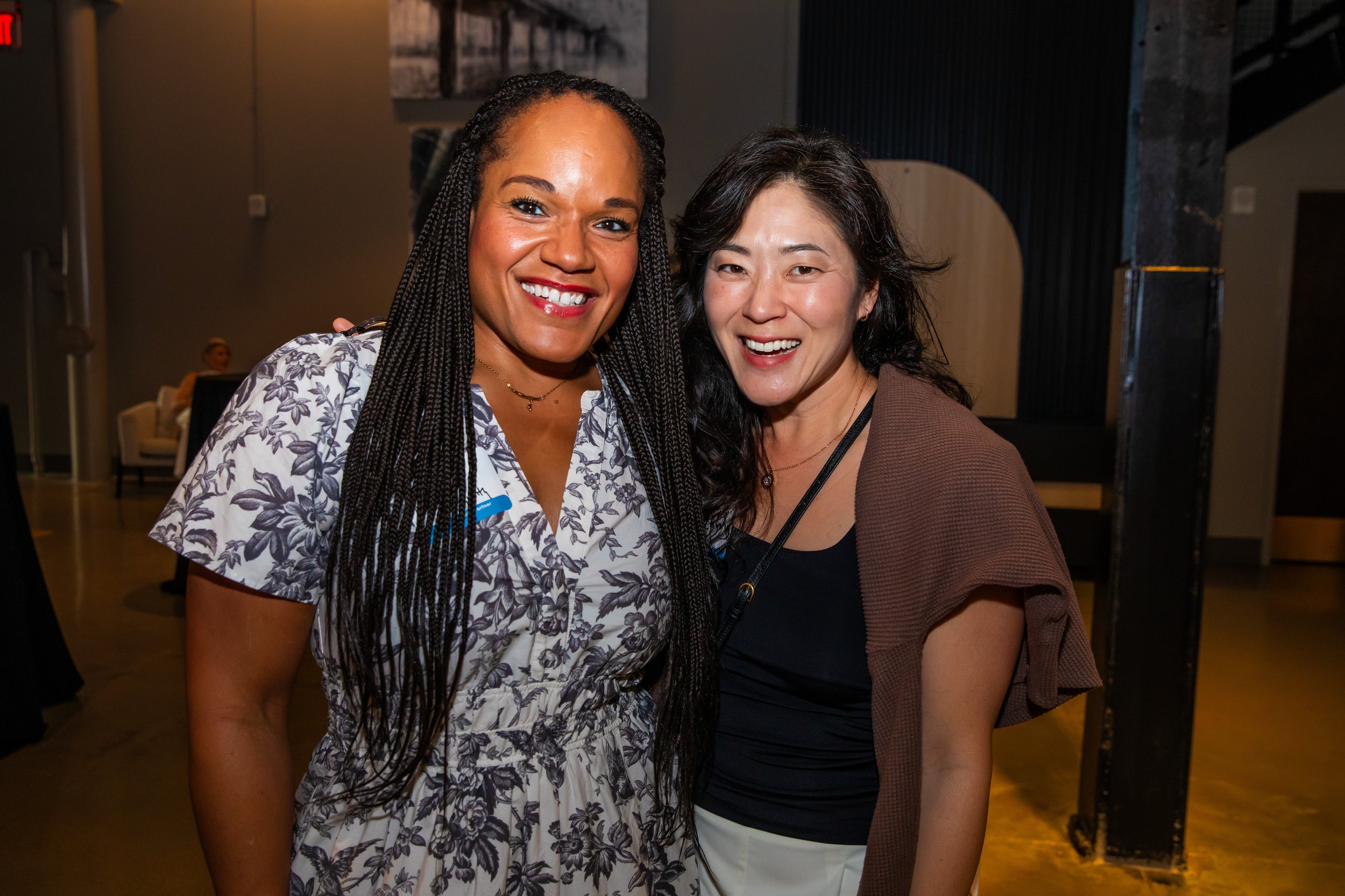 Two women smiling and posing for a photo indoors at a social event. One woman has dark braided hair and the other has dark wavy hair. They are standing close together.
