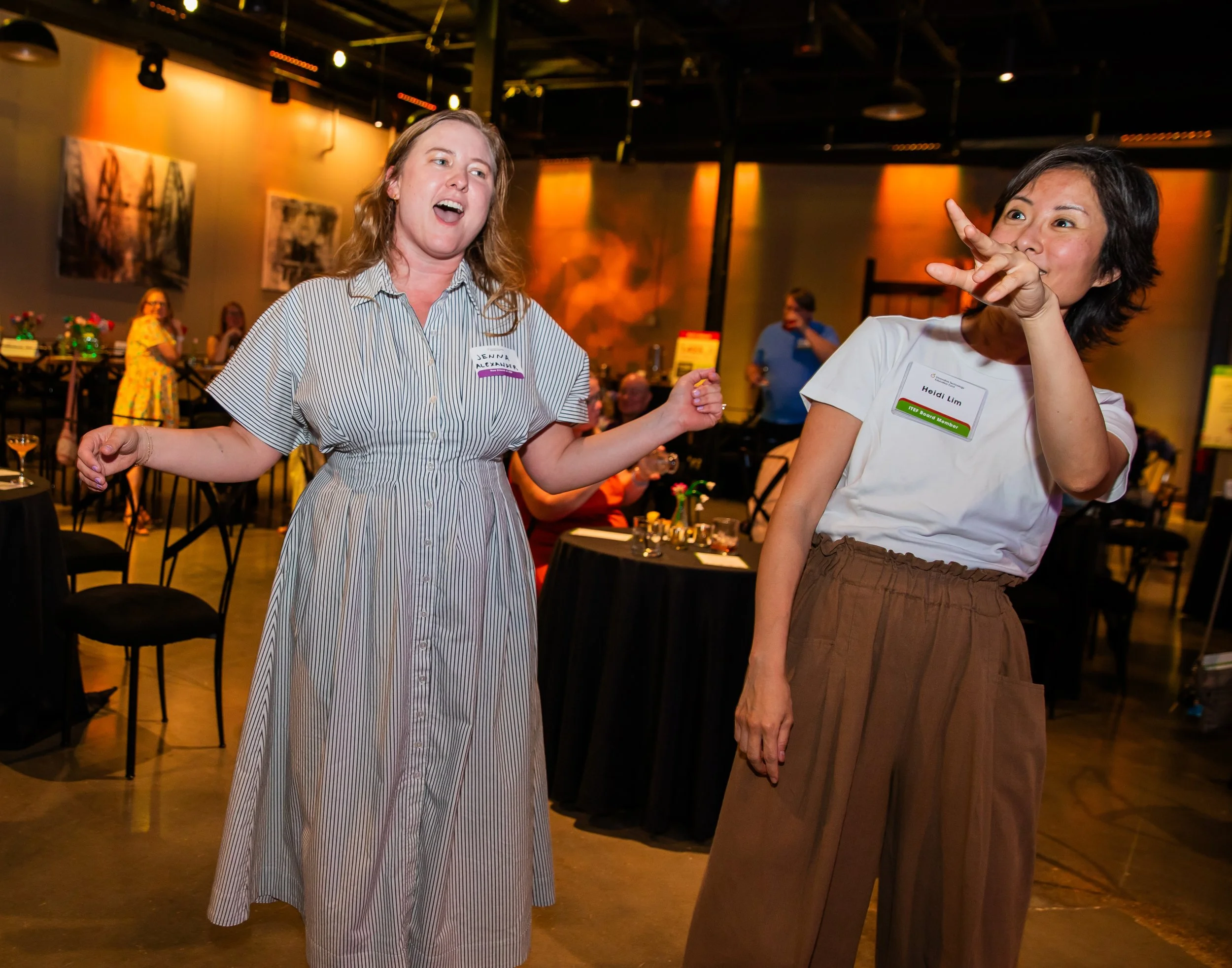 Two women are in conversation at an indoor event. One woman has long, wavy, red hair and is wearing a striped dress, with a name tag that reads 'Jenna Alexander.' The other woman has short, dark hair and is wearing a white top with a green banner on 