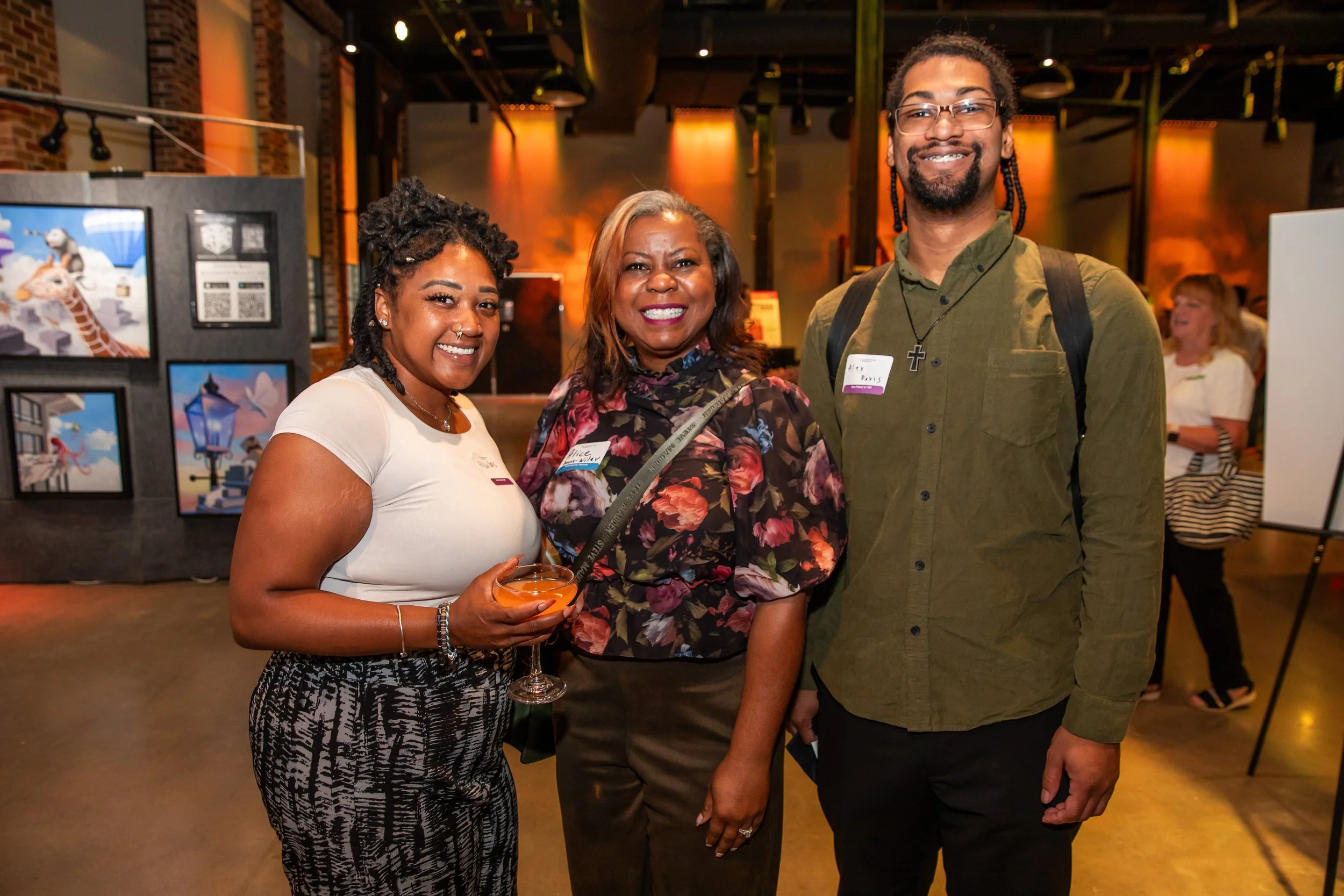 Three people are posing together at an indoor event, smiling; two women and one man. The woman on the left has short curly hair, is holding a cocktail, and is wearing a white top and patterned pants. The woman in the middle has shoulder-length gray h