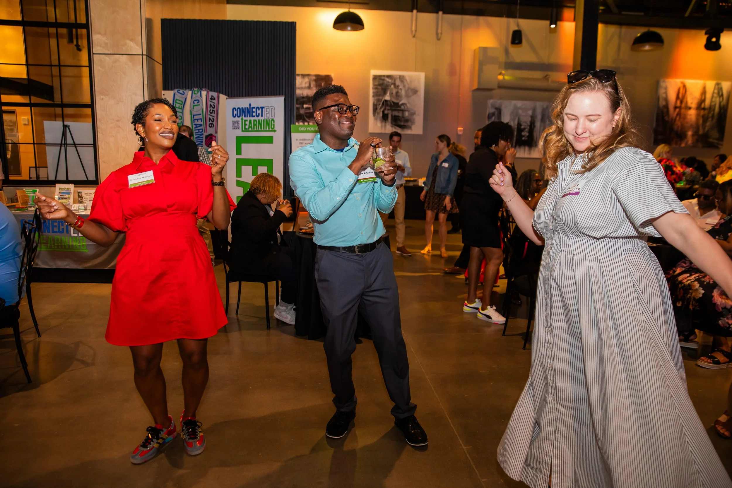 People dancing and socializing at a lively indoor event, with a woman in a red dress and sneakers, a man in glasses wearing a light blue shirt, and a woman in striped attire. There are informational booths and artwork in the background.