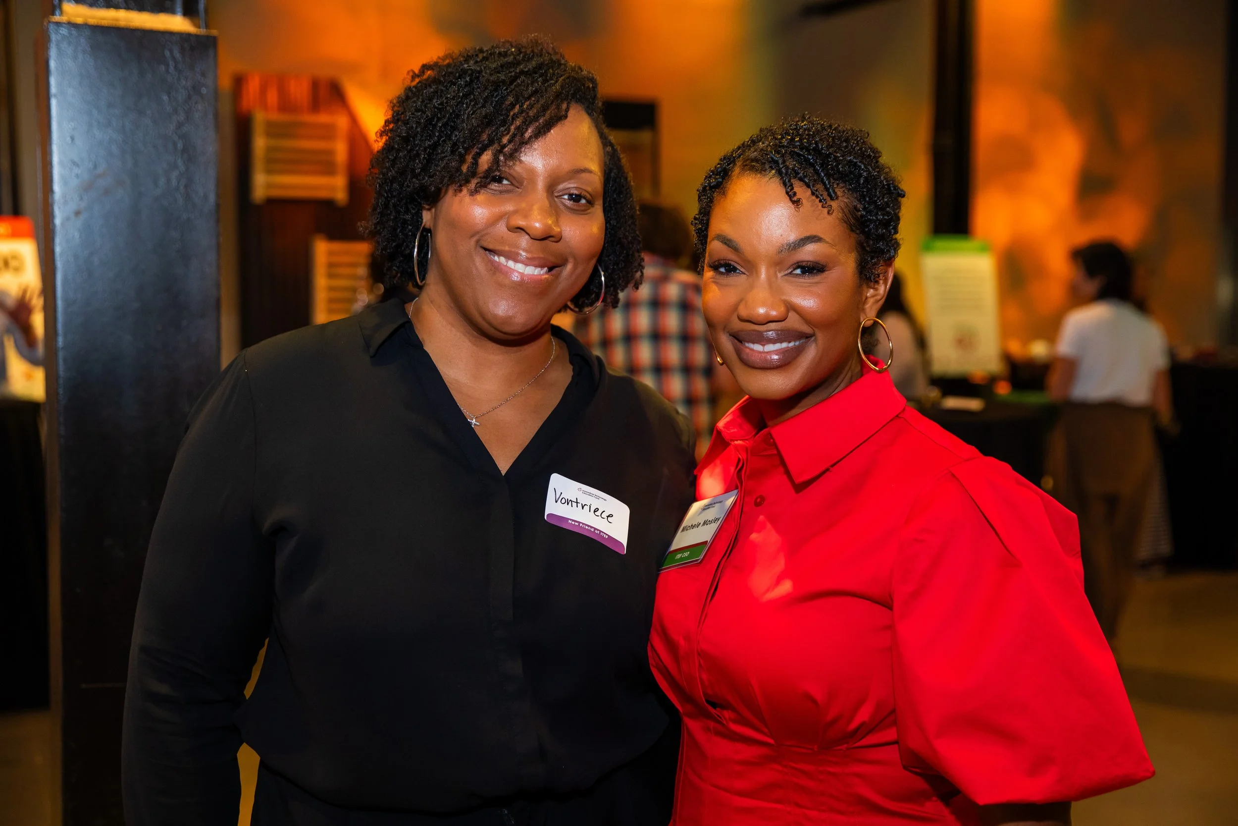 Two women standing close together indoors at what appears to be a social event, smiling at the camera. One woman is wearing a black blouse with a badge that reads 'Vontridge,' and the other woman is wearing a red blouse with a badge.