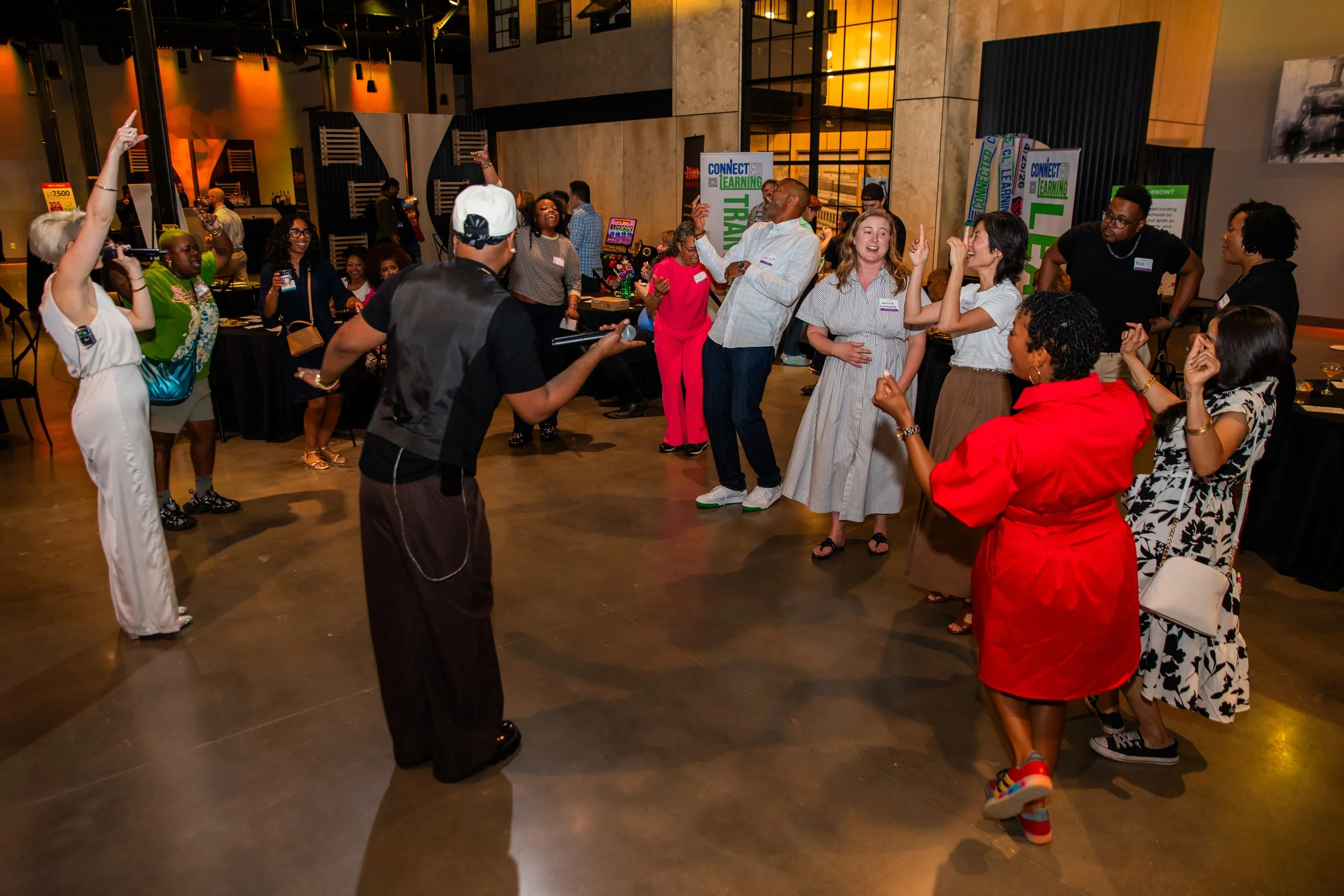 Group of diverse people dancing and enjoying themselves at an indoor event, with some holding microphones and others capturing the moment on their phones, in a modern venue with banners and warm lighting.