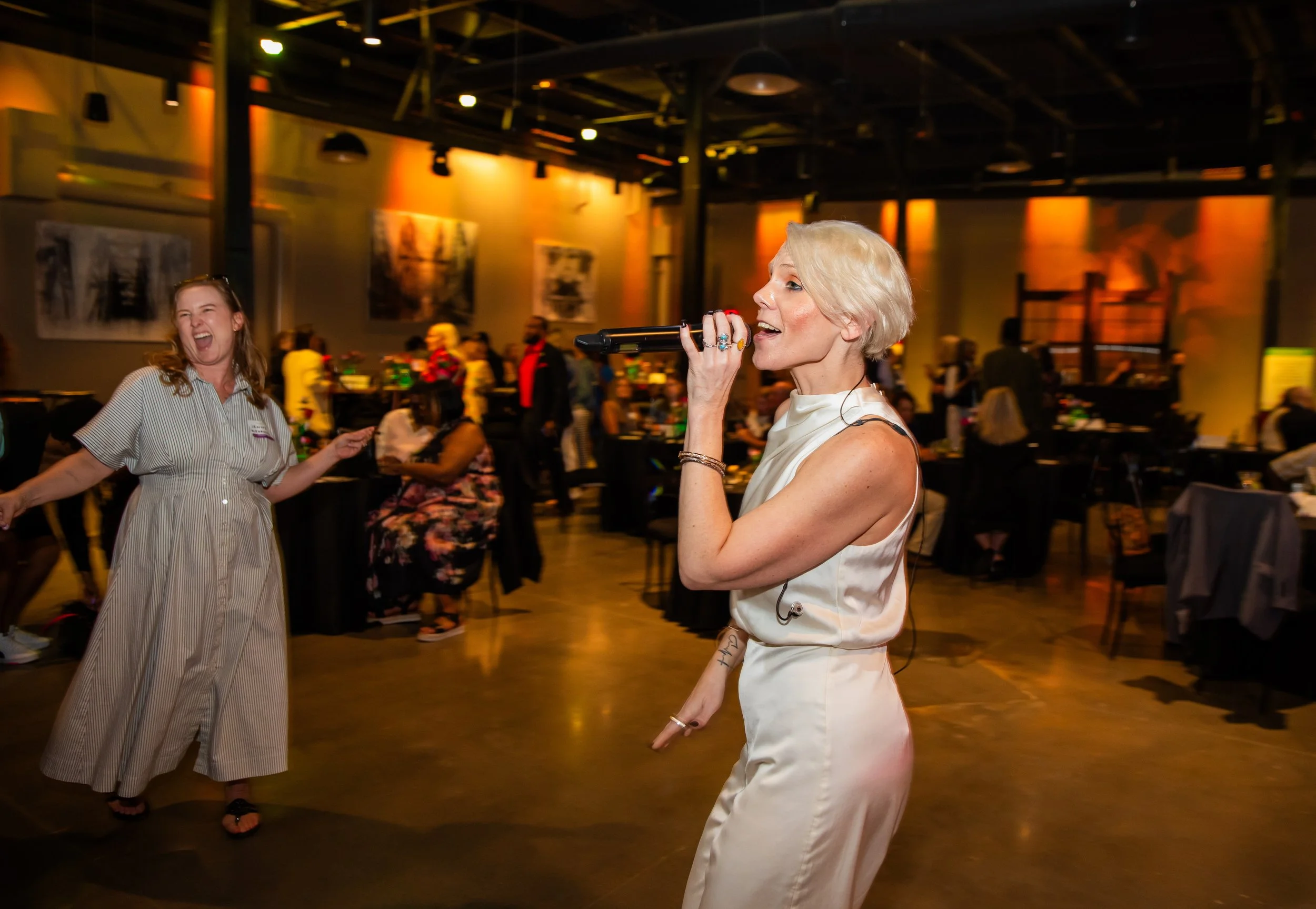 Women singing and dancing at an indoor event with people in the background.