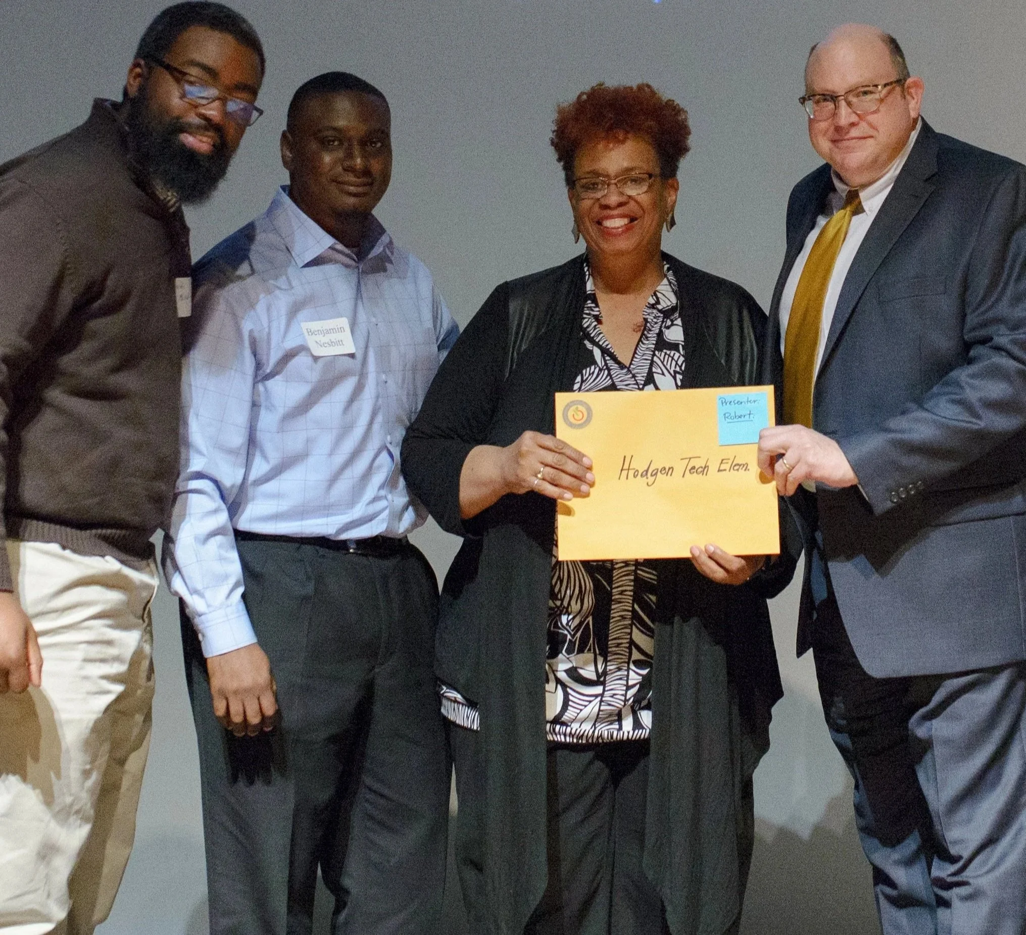 Group of four people standing together at an event, with one holding a yellow sign that has writing on it. The individuals are dressed in business casual attire and are smiling.