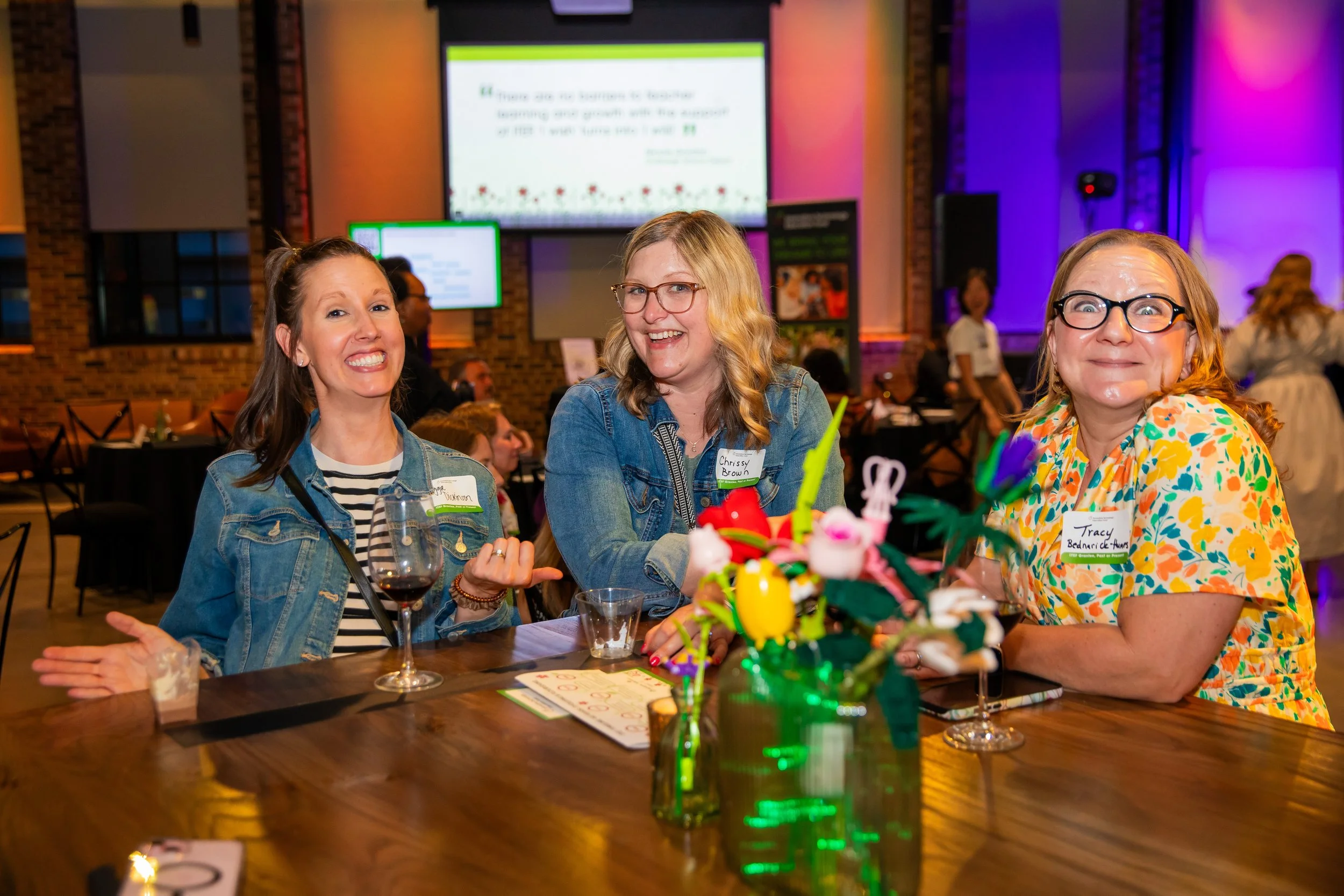 Three women smiling and sitting at a table in a decorated event space with colorful lighting. They are wearing name tags and are surrounded by flowers and drinks.