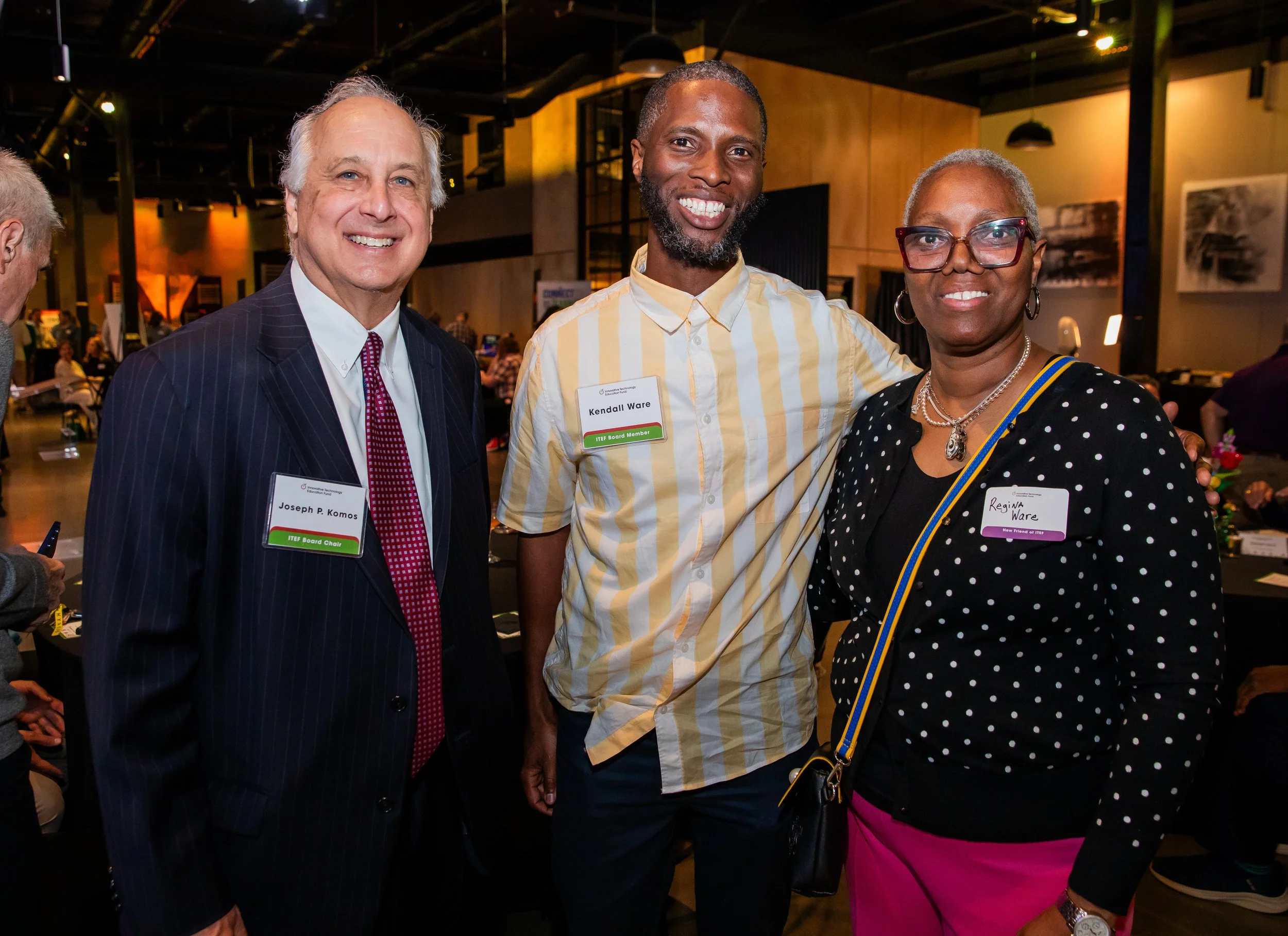 Three people at a social event, smiling at the camera. They are wearing name tags, and the background indicates an indoor venue with warm lighting, other attendees, and some artwork on the walls.