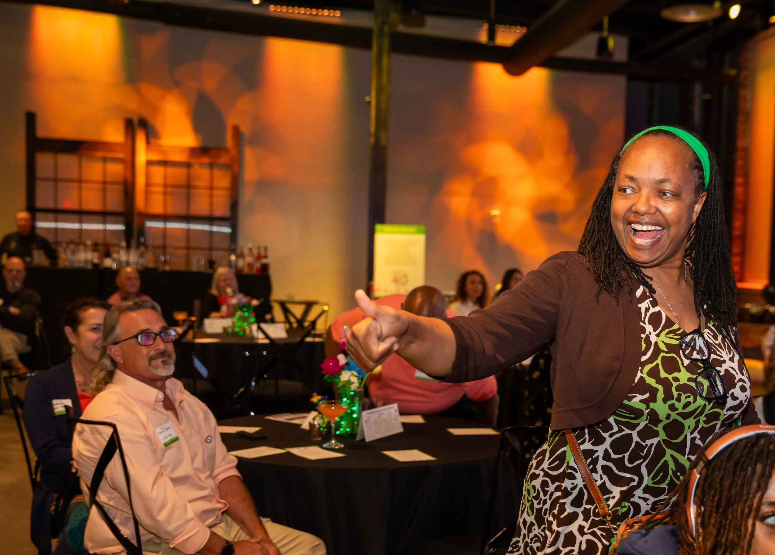 A woman with dark braided hair and a green headband is smiling and enthusiastically interacting at a social event. She is wearing a dress with a black, white, and green abstract pattern and sunglasses hanging around her neck. The background shows peo