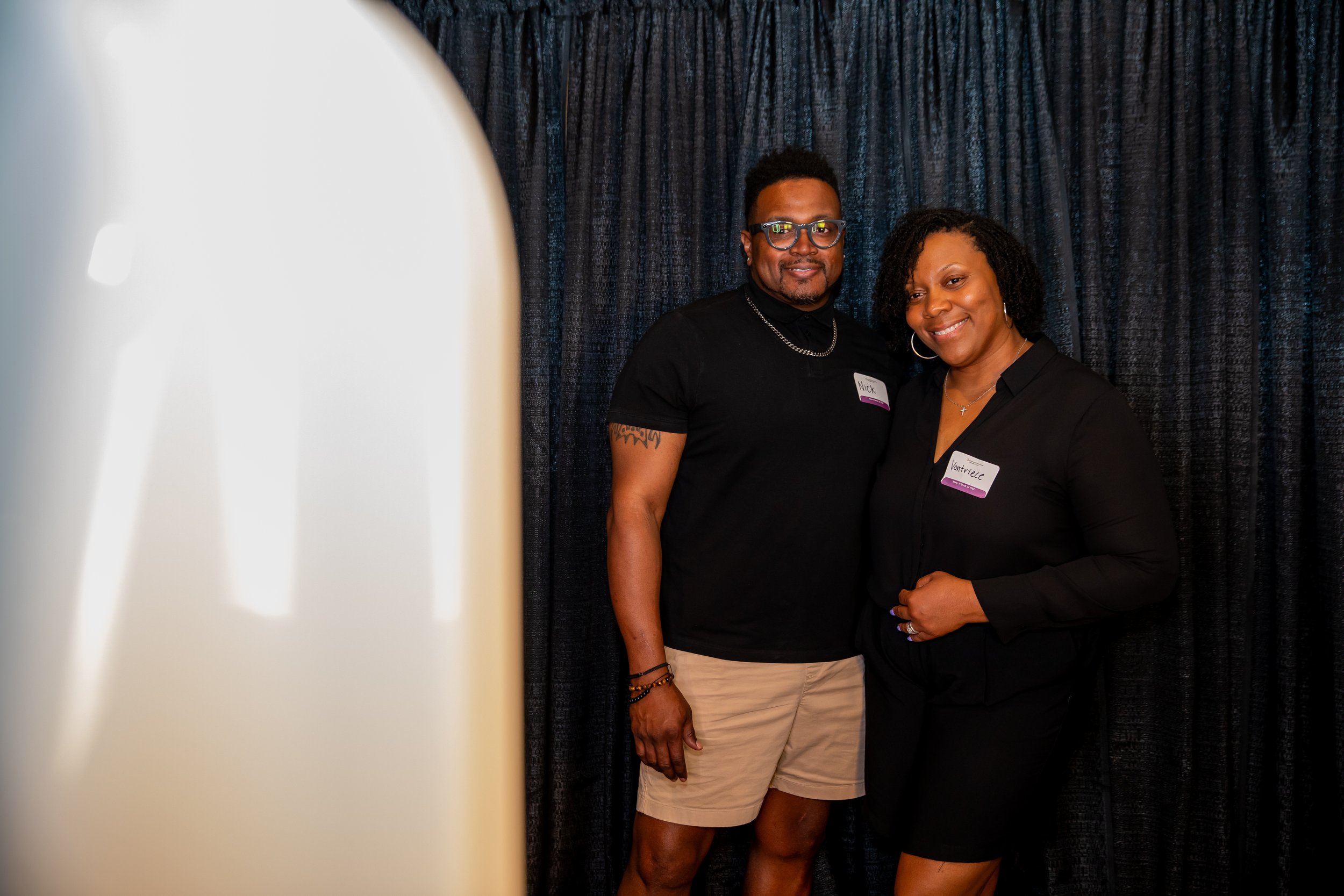 Two people standing in front of a dark curtain, smiling at the camera. One person is a man wearing glasses and a black shirt with a name tag that says "Nick." The other is a woman with curly hair wearing a black top, also with a name tag that says "V