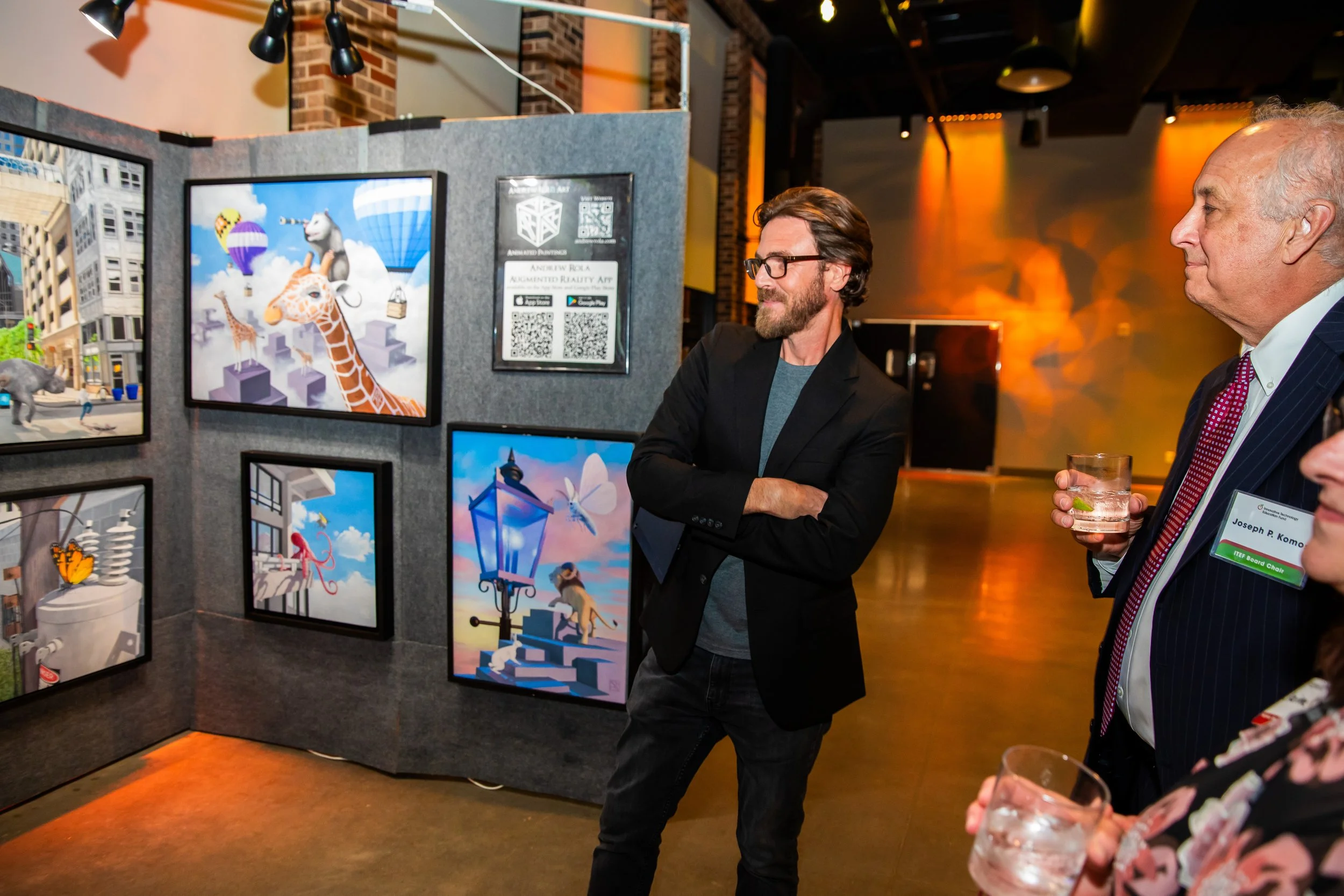 Two men standing in front of a display board at an art exhibit, looking at digital artworks depicting animals and city scenes. One man with glasses and a beard has his arms crossed, and the other man is holding a glass of drink.