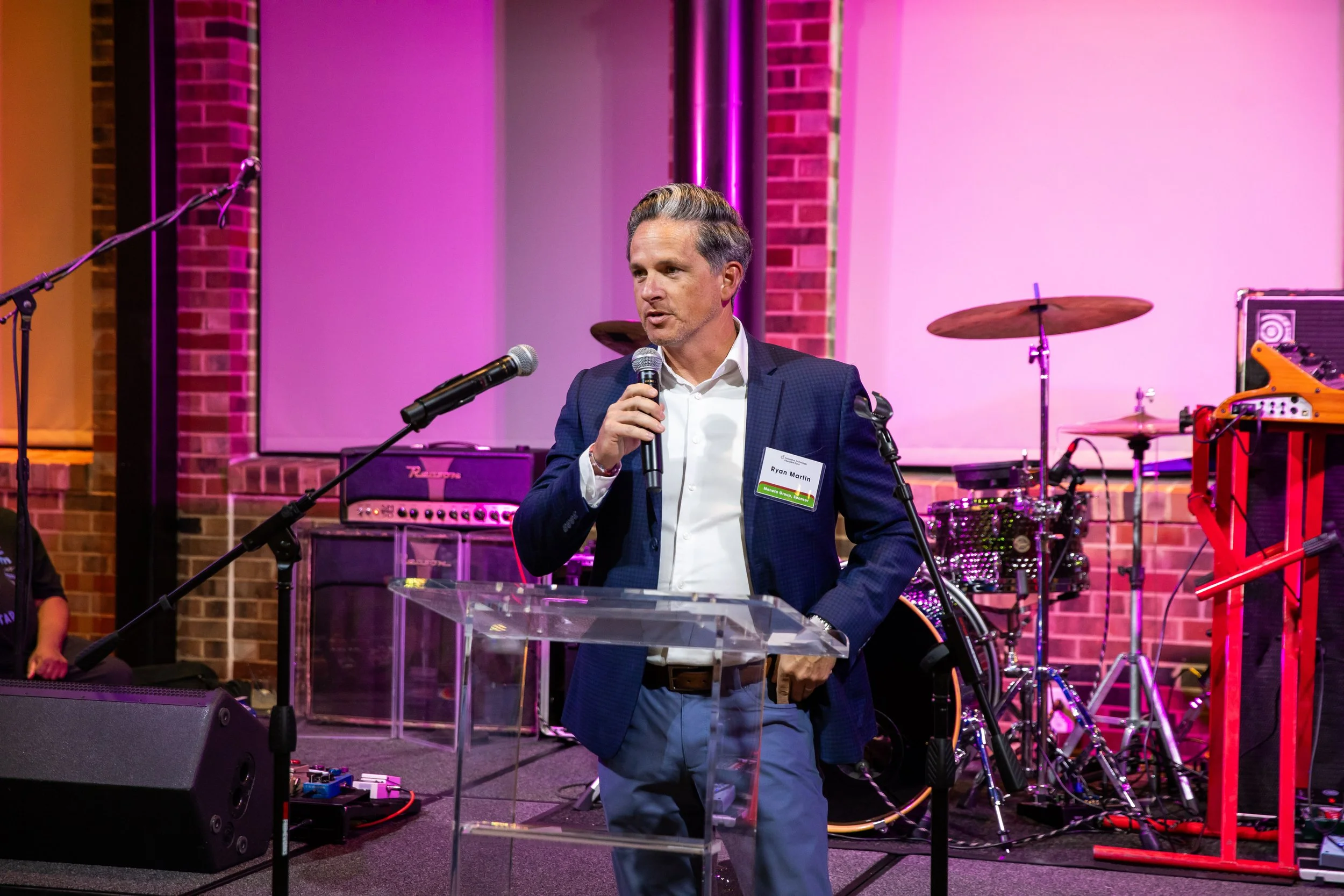 A man in a dark suit gives a speech at a podium with a microphone, in a venue with pink lighting and musical instruments behind him.