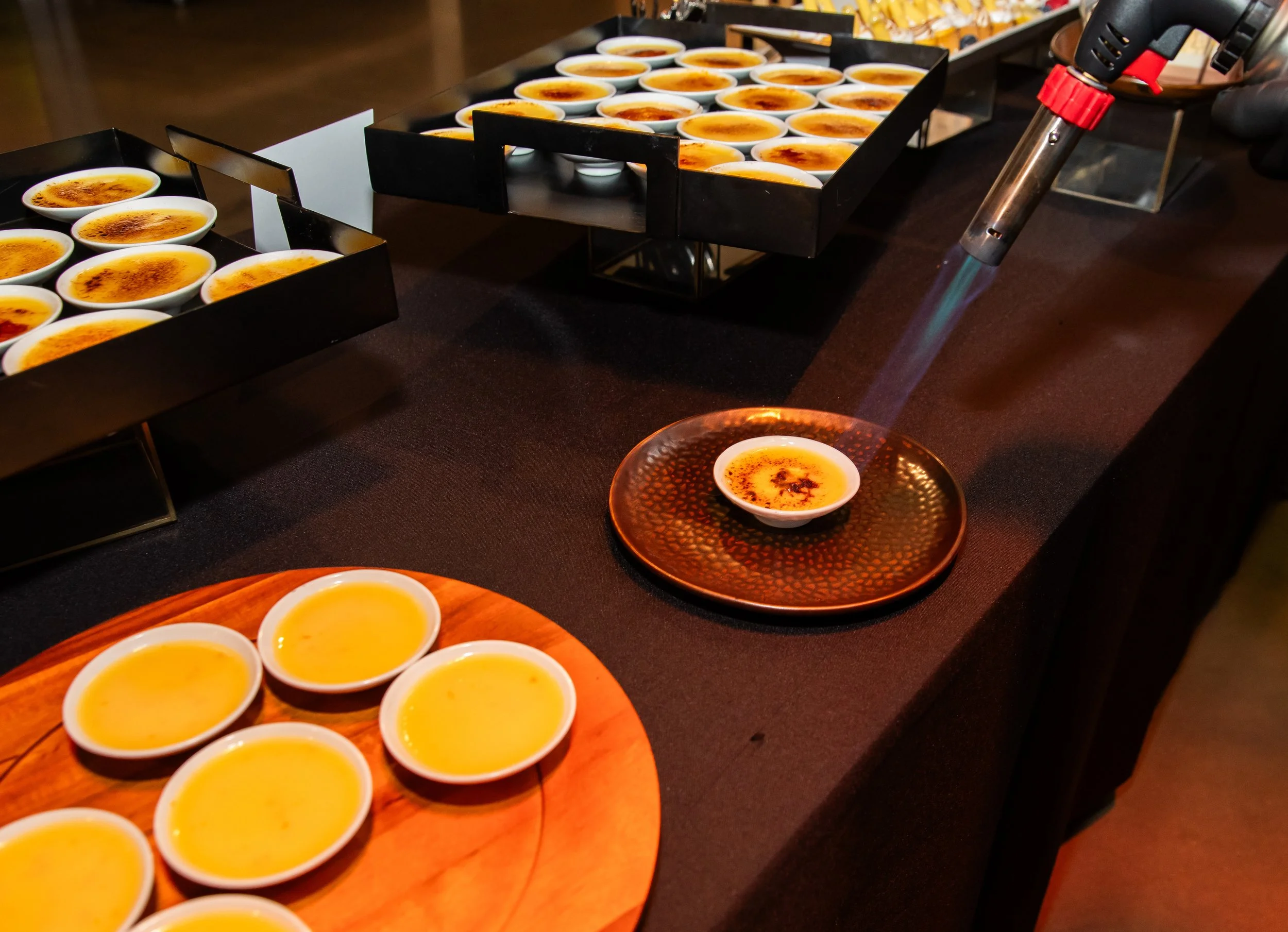 A table with several small white bowls filled with a yellow dessert, possibly crème brûlée, some with a caramelized sugar topping. There are also black trays holding more bowls and a culinary torch used for caramelizing the sugar.