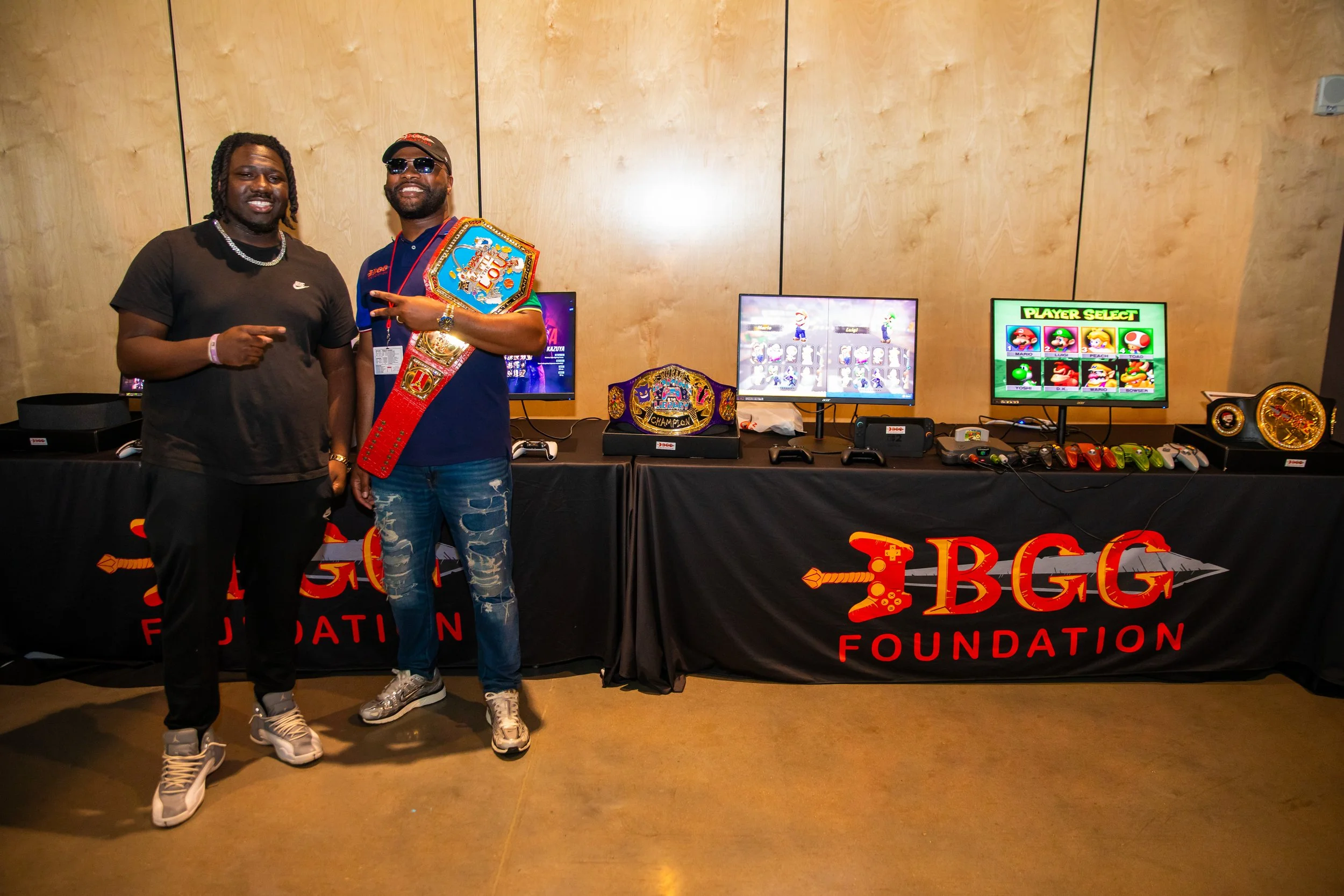 Two men standing in front of a gaming setup at the BGG Foundation event. One man is pointing at the other, who is wearing sunglasses and holding a championship belt and a large gaming trophy. The background features monitors displaying video game cha