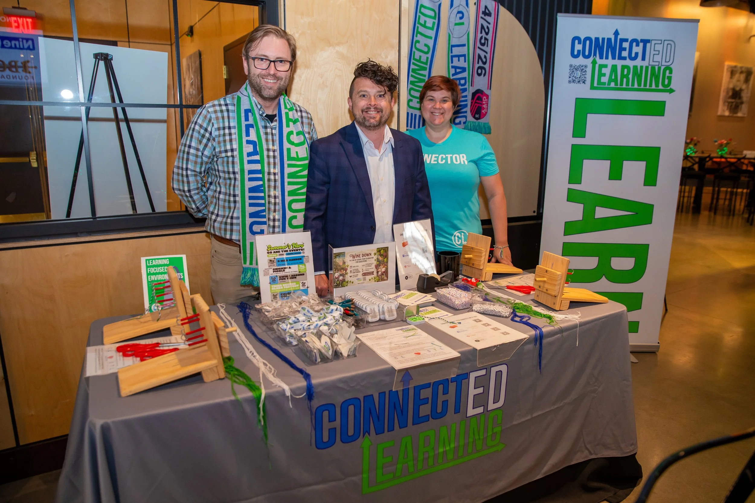 A group of three smiling people standing behind a table with educational materials and small wooden displays at a CONNECTED LEARNING event, with a large banner and colorful signs in the background.