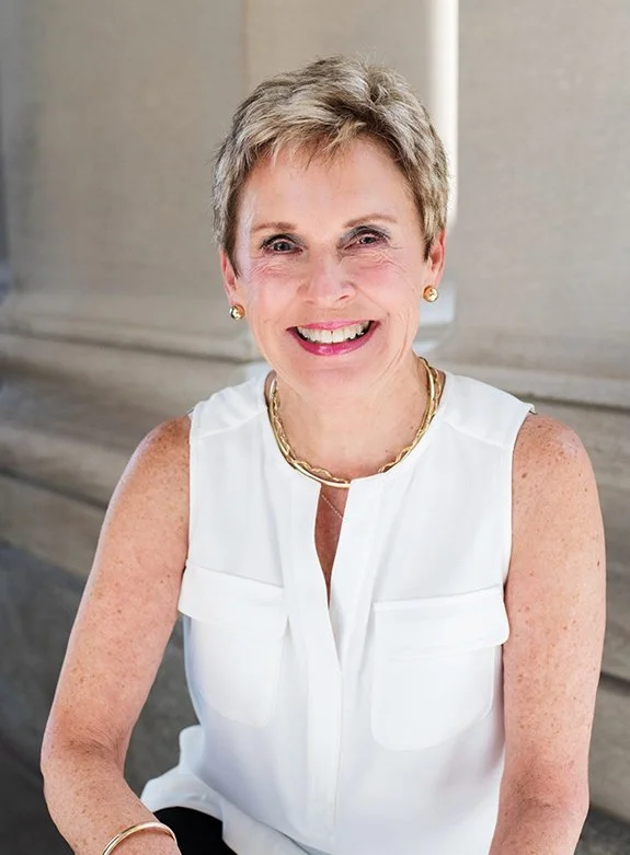 A smiling middle-aged woman with short gray hair, wearing a white sleeveless blouse, gold jewelry, and sitting outdoors in front of a beige stone building.