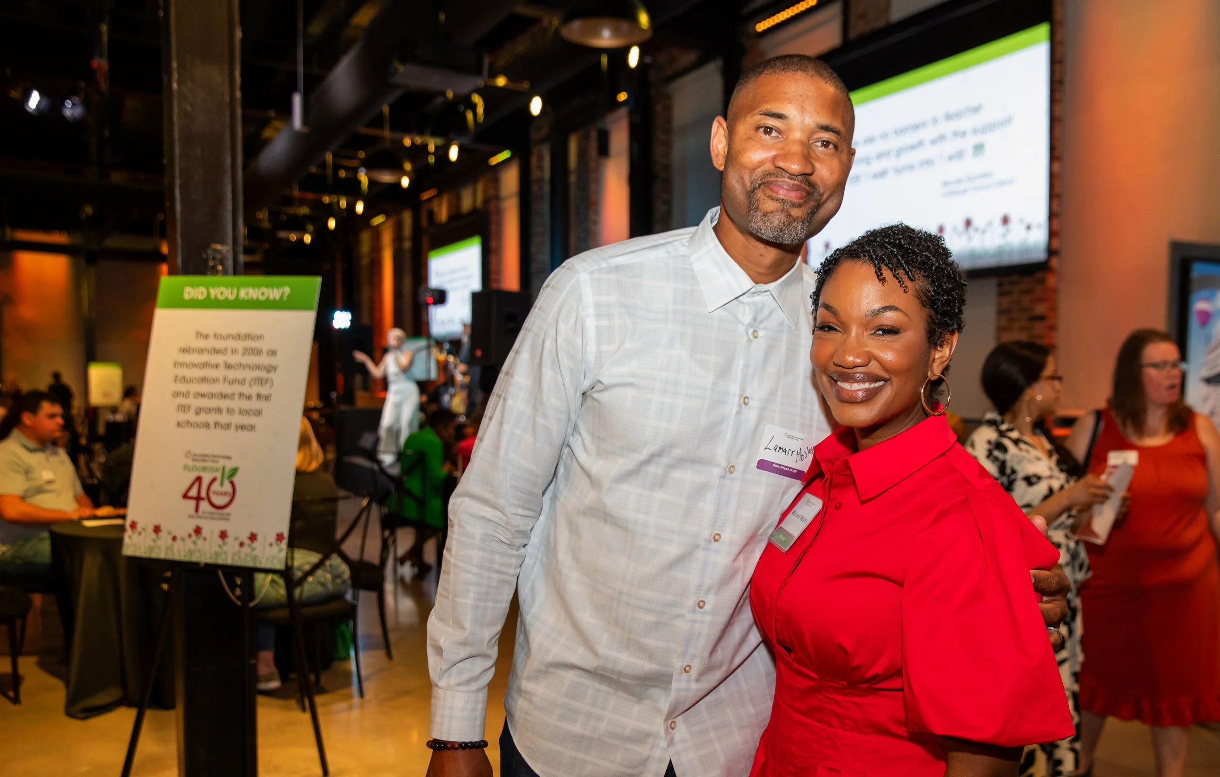 A man and woman are smiling and posing together at an indoor event, with large screens and other attendees in the background.