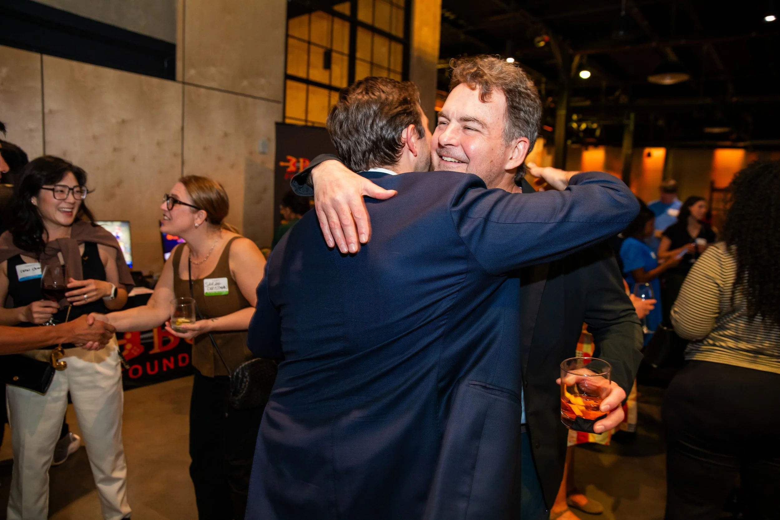 Two men hugging at a social event, smiling. Other people standing around, some holding drinks, in a warm, lively indoor setting with dim lighting.