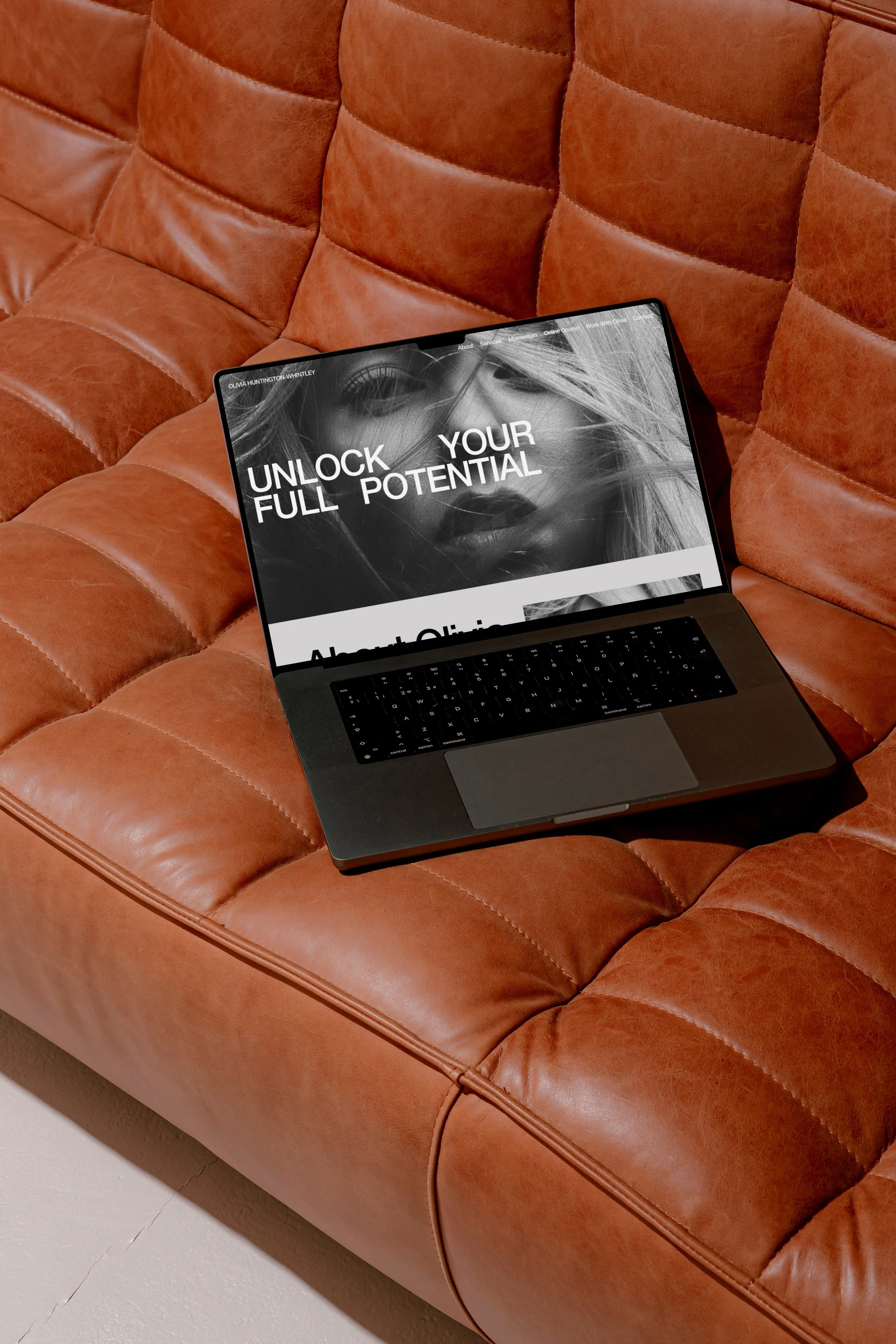 A laptop with a promotional website on its screen, resting on a tufted, brown leather sofa.