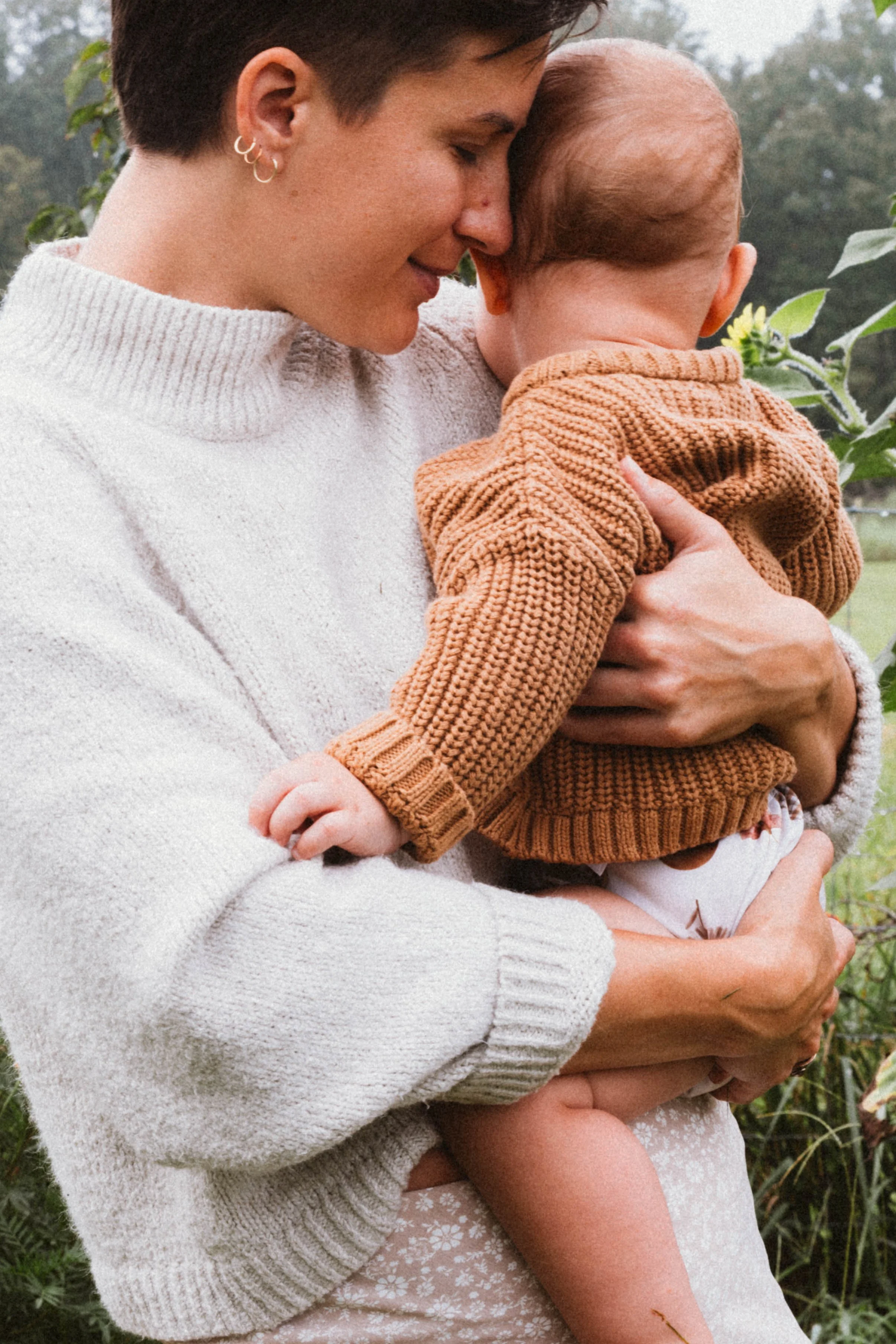 A woman holding a baby outdoors, close to a sunflower plant, with lush green trees in the background. The woman is wearing a cream-colored sweater and has short, dark hair, while the baby is dressed in a brown knit sweater.