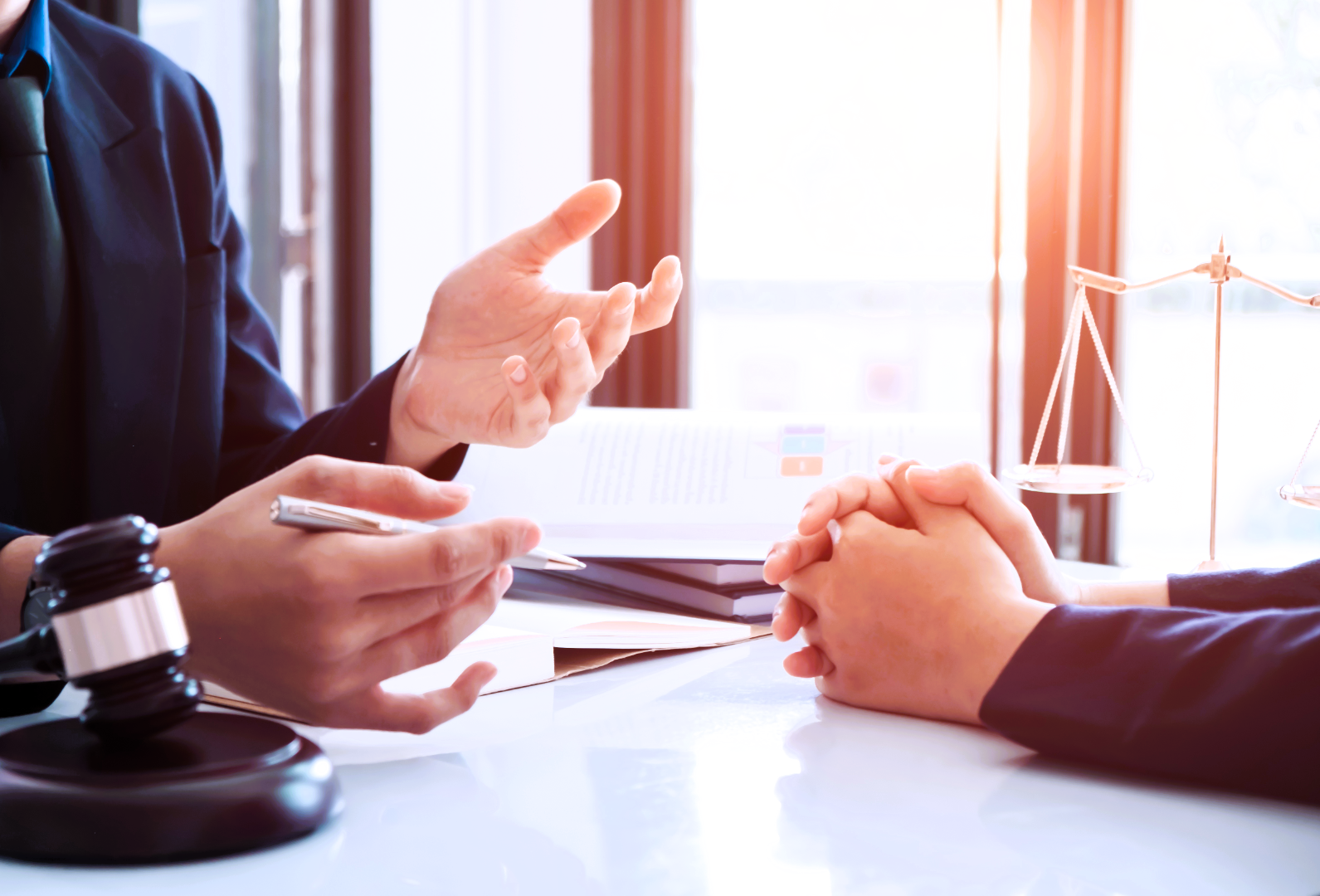 A doctor consulting with a patient, with the doctor gesturing and the patient clasping hands on the table. A stethoscope and a scale model of scales of justice are on the desk, near a window.