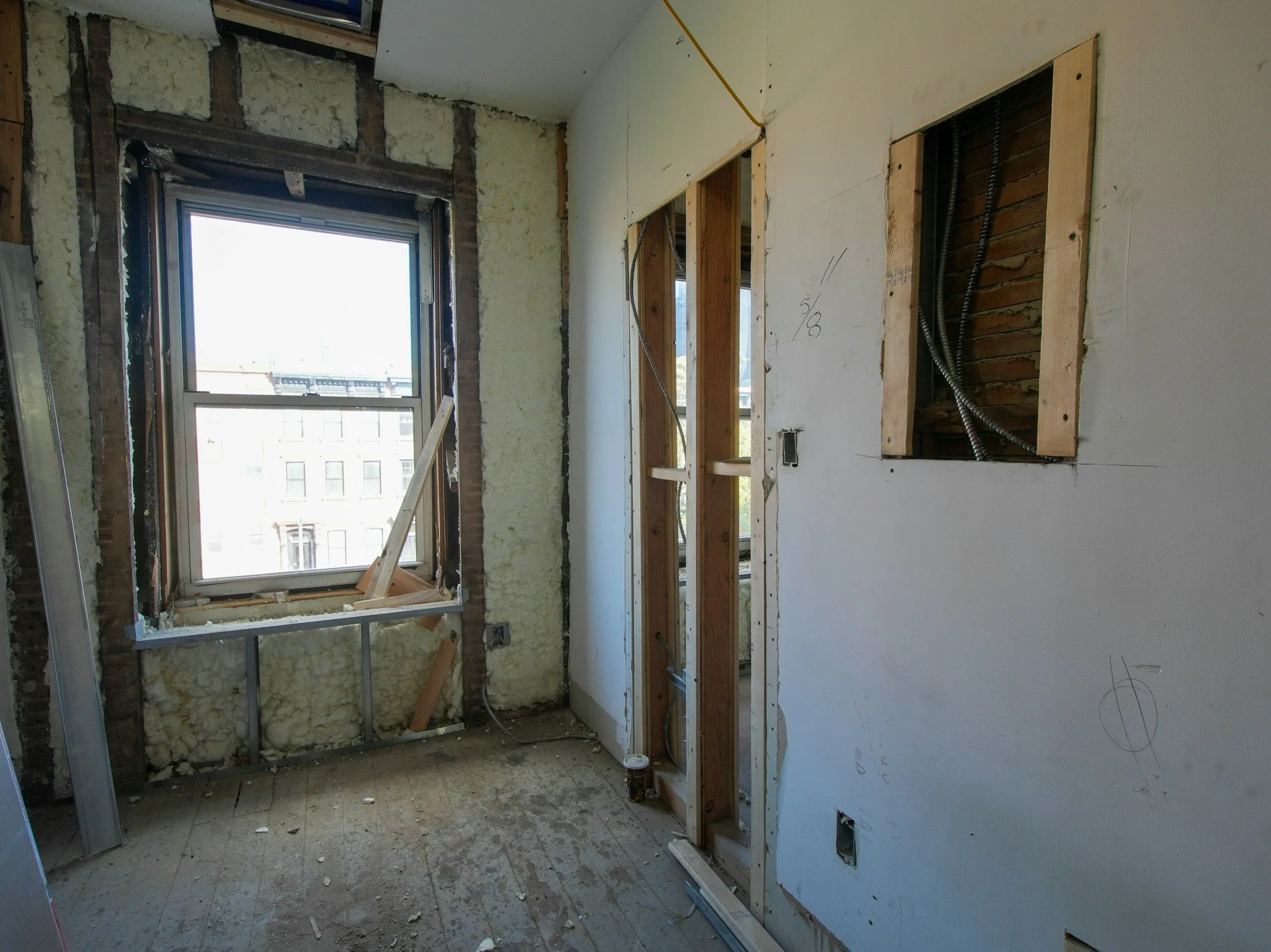 Interior of a room under renovation showing a window, exposed wall studs, some electrical wiring, and construction materials.