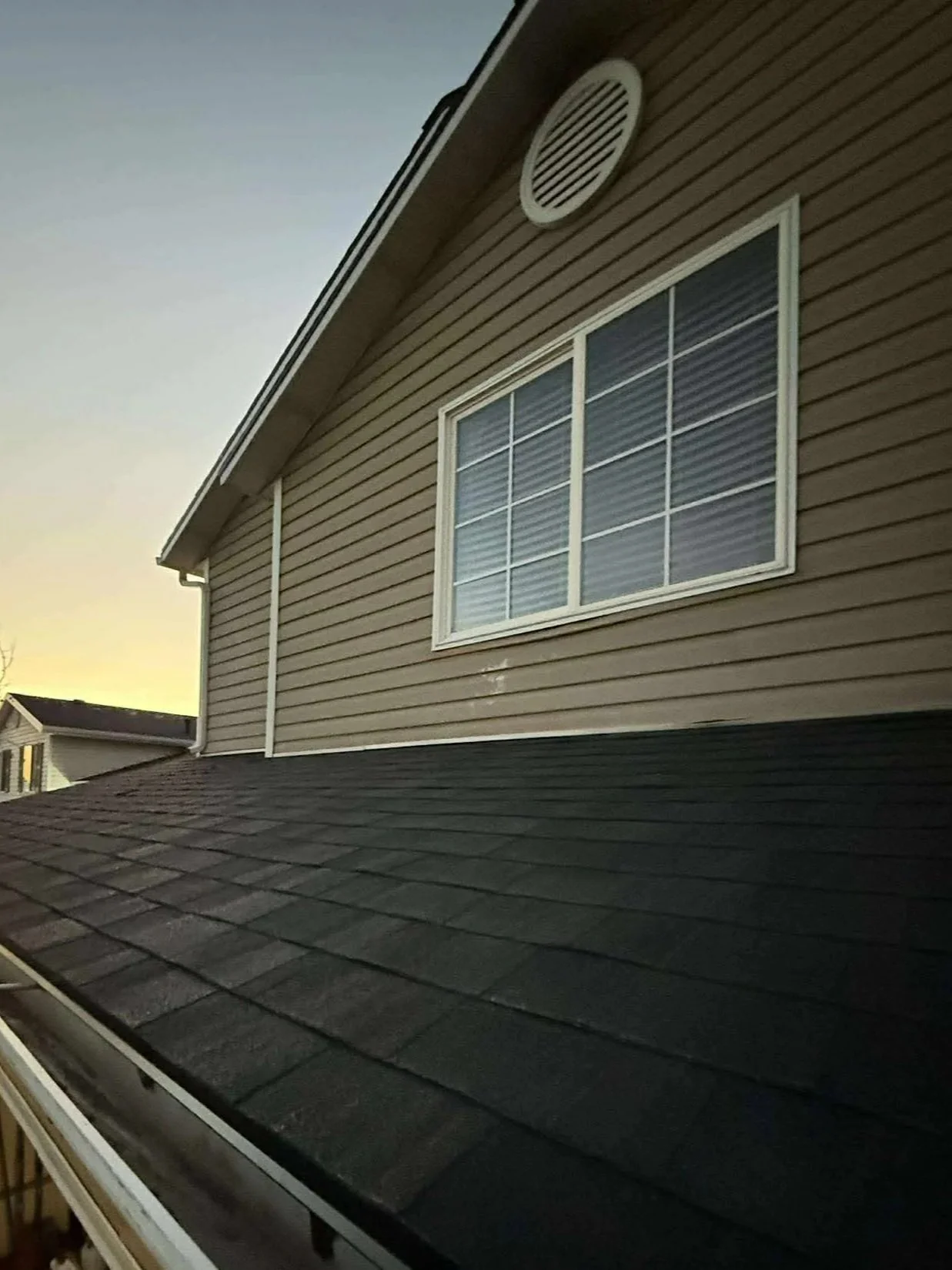 The image shows a house with beige vinyl siding, a large window with white trim, and a vent near the roof's peak at sunset.