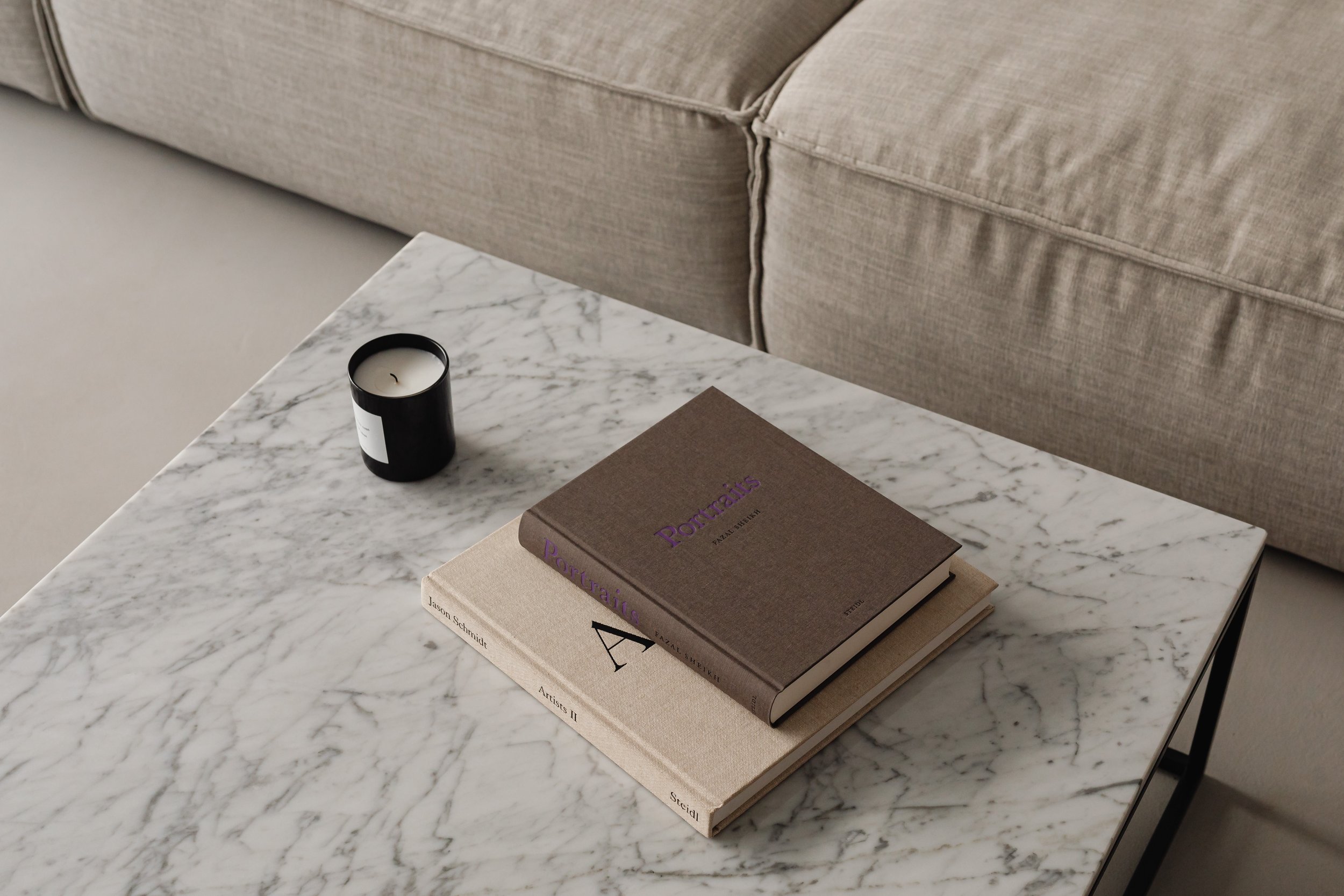 A marble coffee table with a black candle and two books stacked, with a beige sofa in the background.