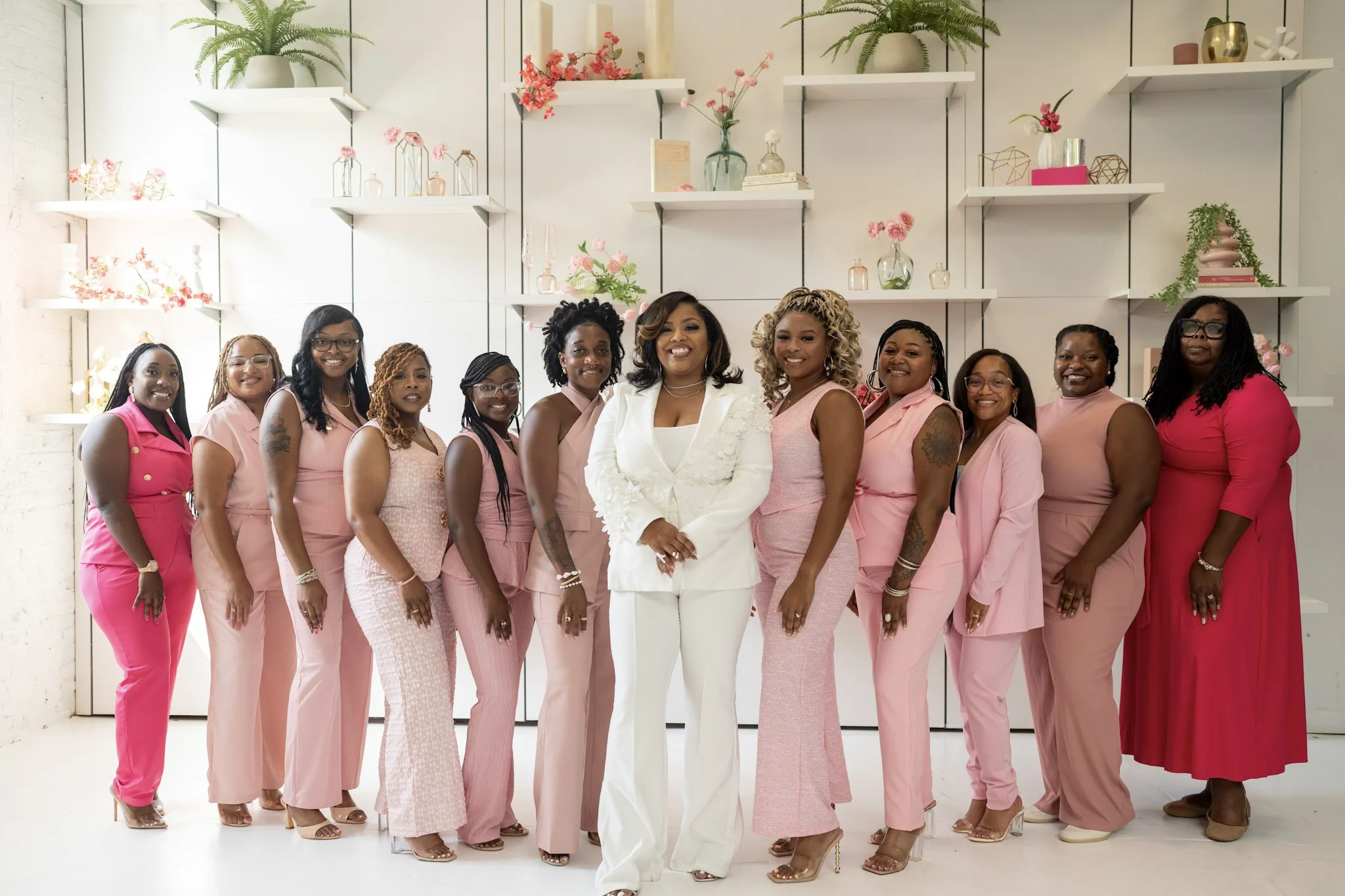 Group of women in pink and white outfits standing together indoors with modern decor and floral arrangements in the background.