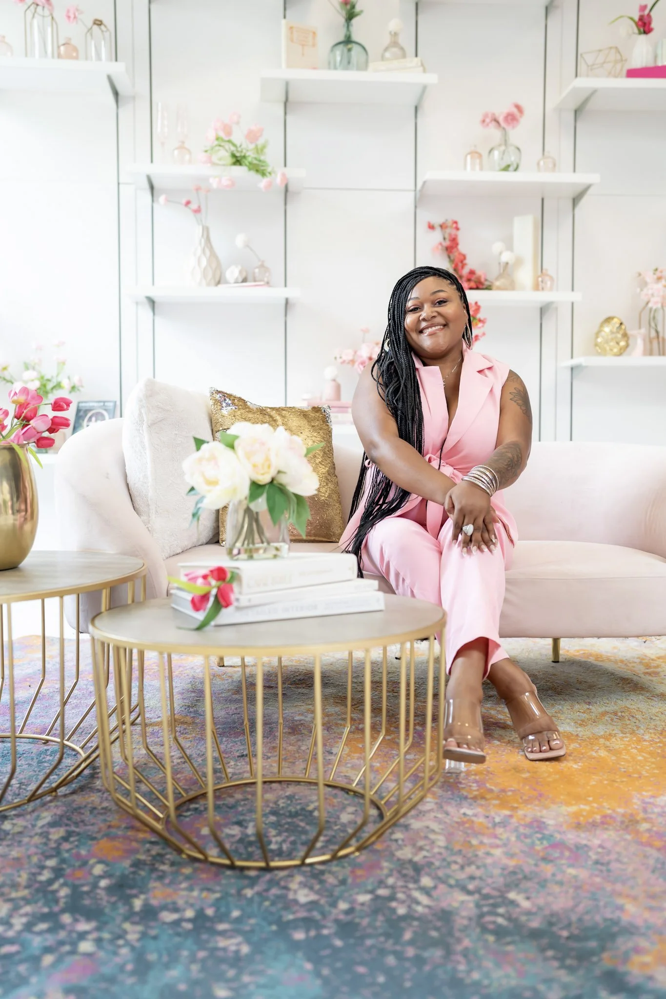 Woman sitting on a pink sofa in a bright, decorated room with vases, flowers, and decorative objects on white shelves, wearing a pink jumpsuit and smiling.