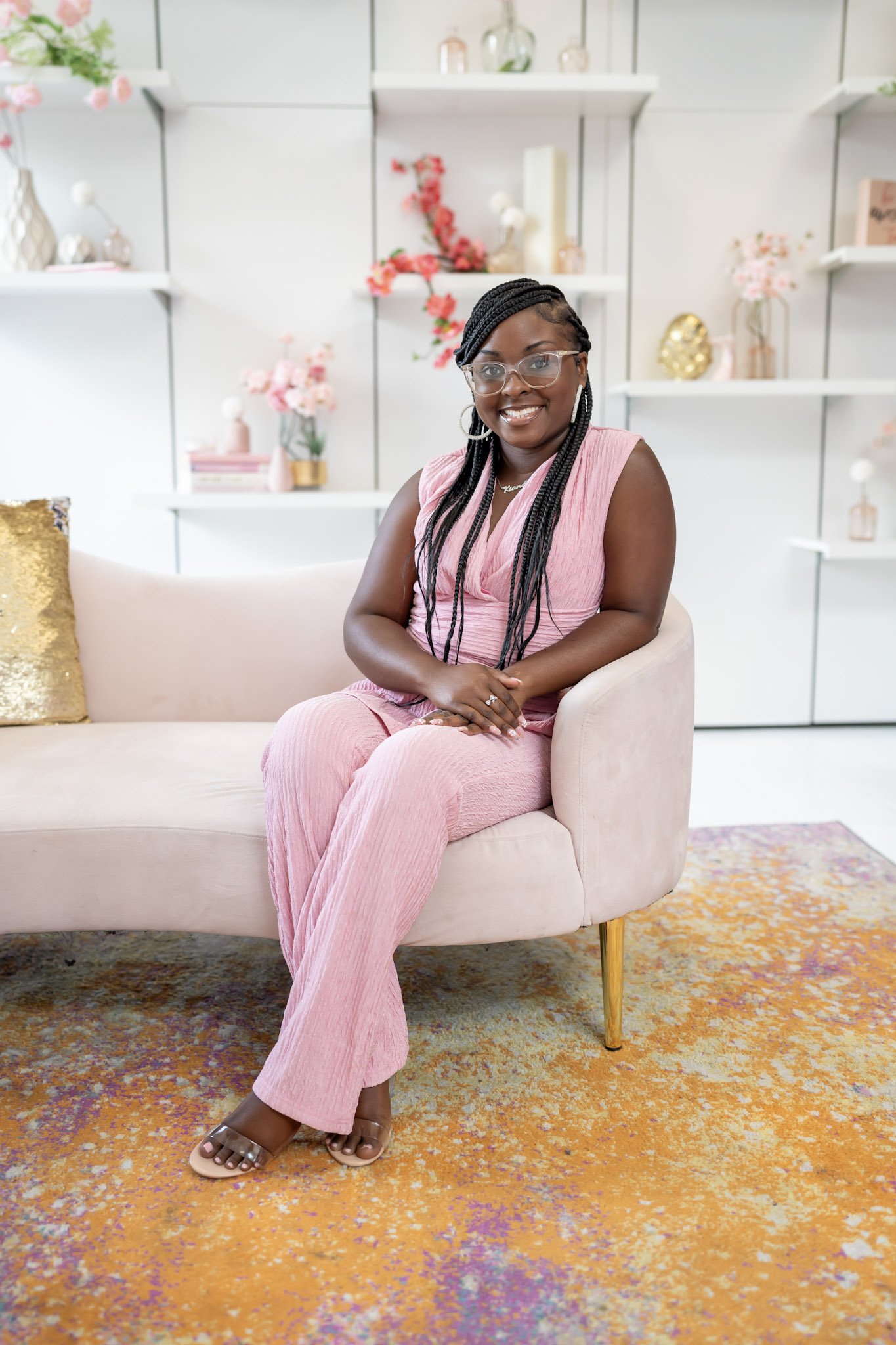 A woman with braided hair and glasses, wearing a pink sleeveless top and pink pants, sitting on a light pink sofa in a decorated room with shelves filled with vases and flowers, and a colorful rug.