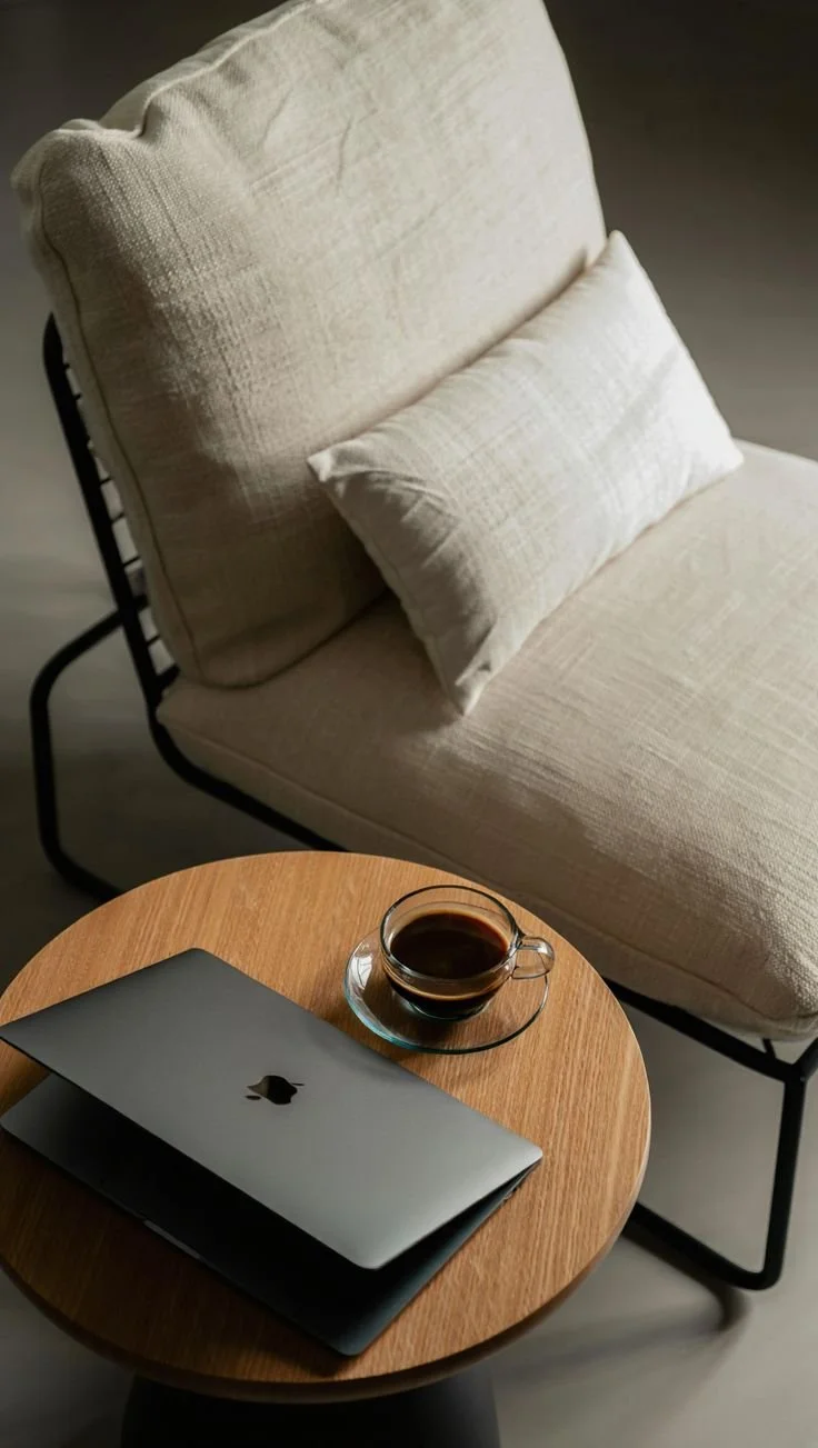 A cozy living space with a beige armchair, a pillow, a small wooden side table, a laptop, and a cup of coffee.