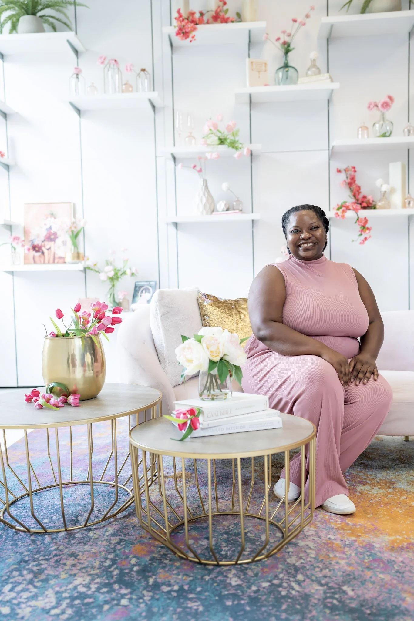 A woman in a pink outfit smiling while sitting on a sofa in a bright, decorated room with pink and white flowers, modern vases, and art on shelves and table.