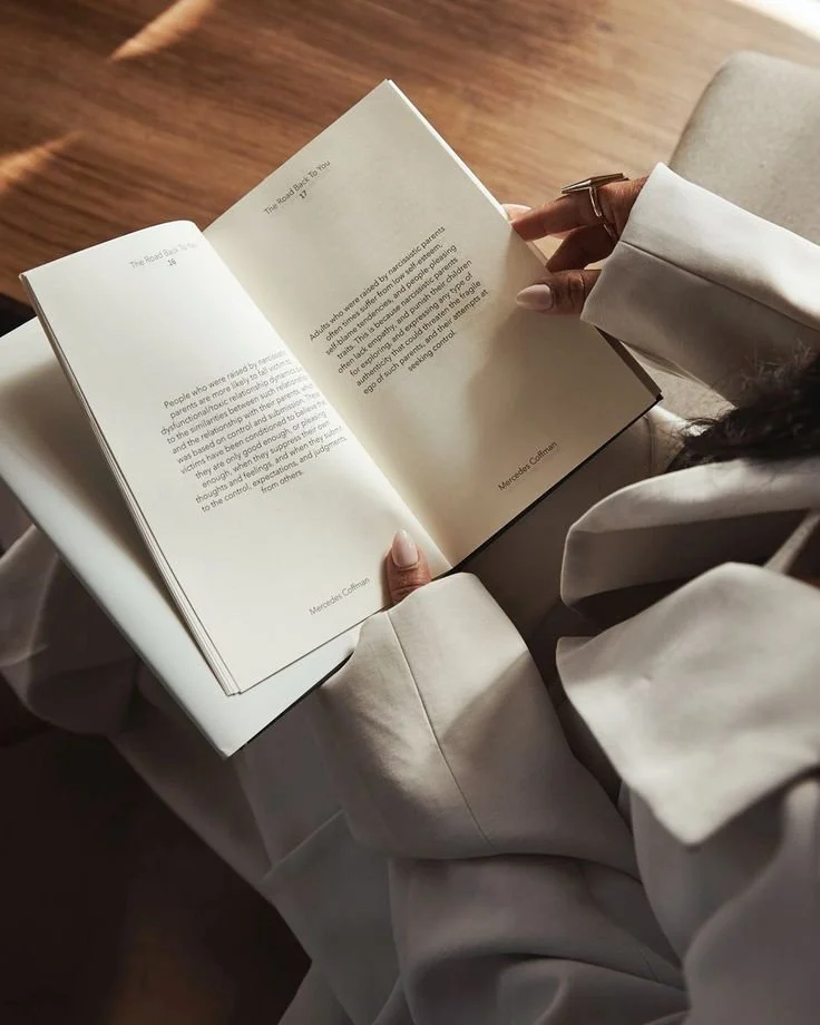 A person wearing a white long-sleeve shirt is holding an open book with white pages. The person is sitting, and the photo is taken from above, focusing on their hands and the book. The background shows a wooden surface and a beige sofa.