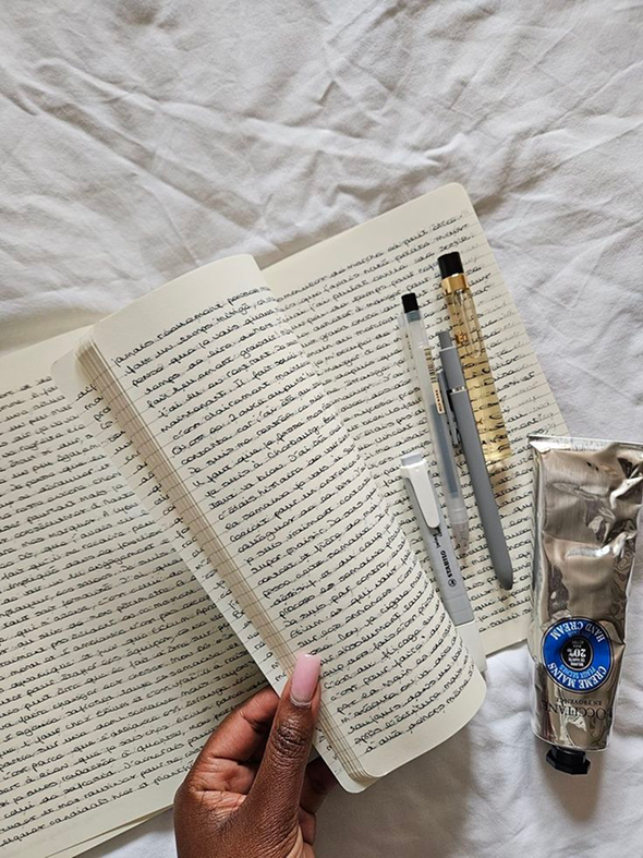 Open notebook with handwritten notes, various pens, a rolled-up sticky note, a tube of perfume, and a crumpled silver foil packet on a white fabric surface.
