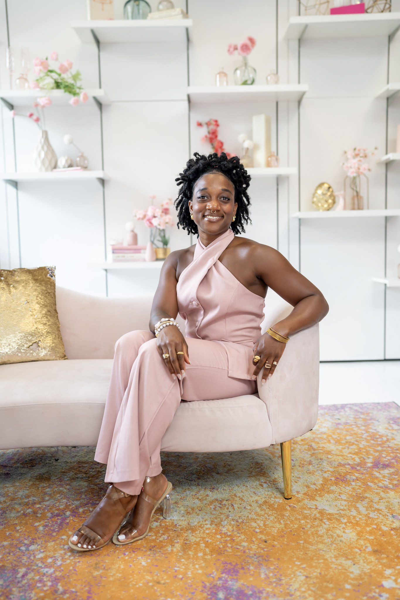 A woman with short curly hair, dark skin, wearing a pink halter jumpsuit, sitting on a light pink sofa in a room decorated with pink and gold accents, flowers, and decorative objects.