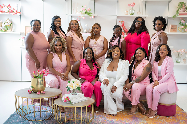 A group of ten women gathered in a bright, floral-themed room. They are dressed in pink and white outfits, seated and standing around three round tables decorated with flowers and books. The women are smiling and appear joyful, celebrating an occasion.