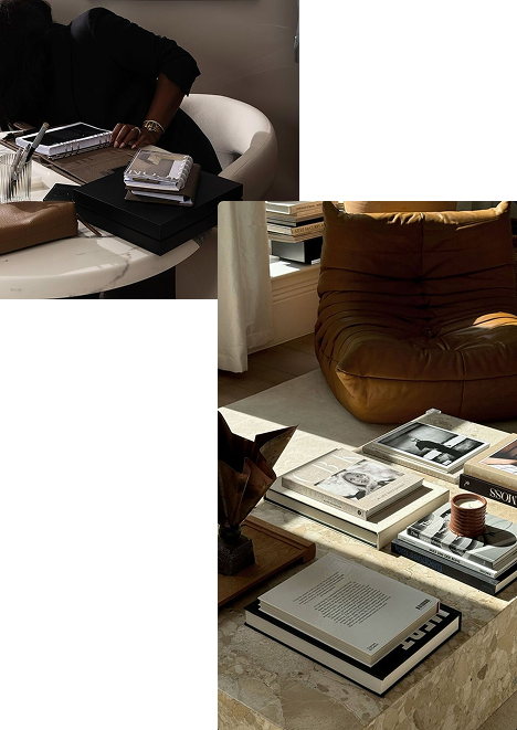 A living room with a brown leather armchair, a stack of books on a table, and a person sitting at a table with more books and a notebook.