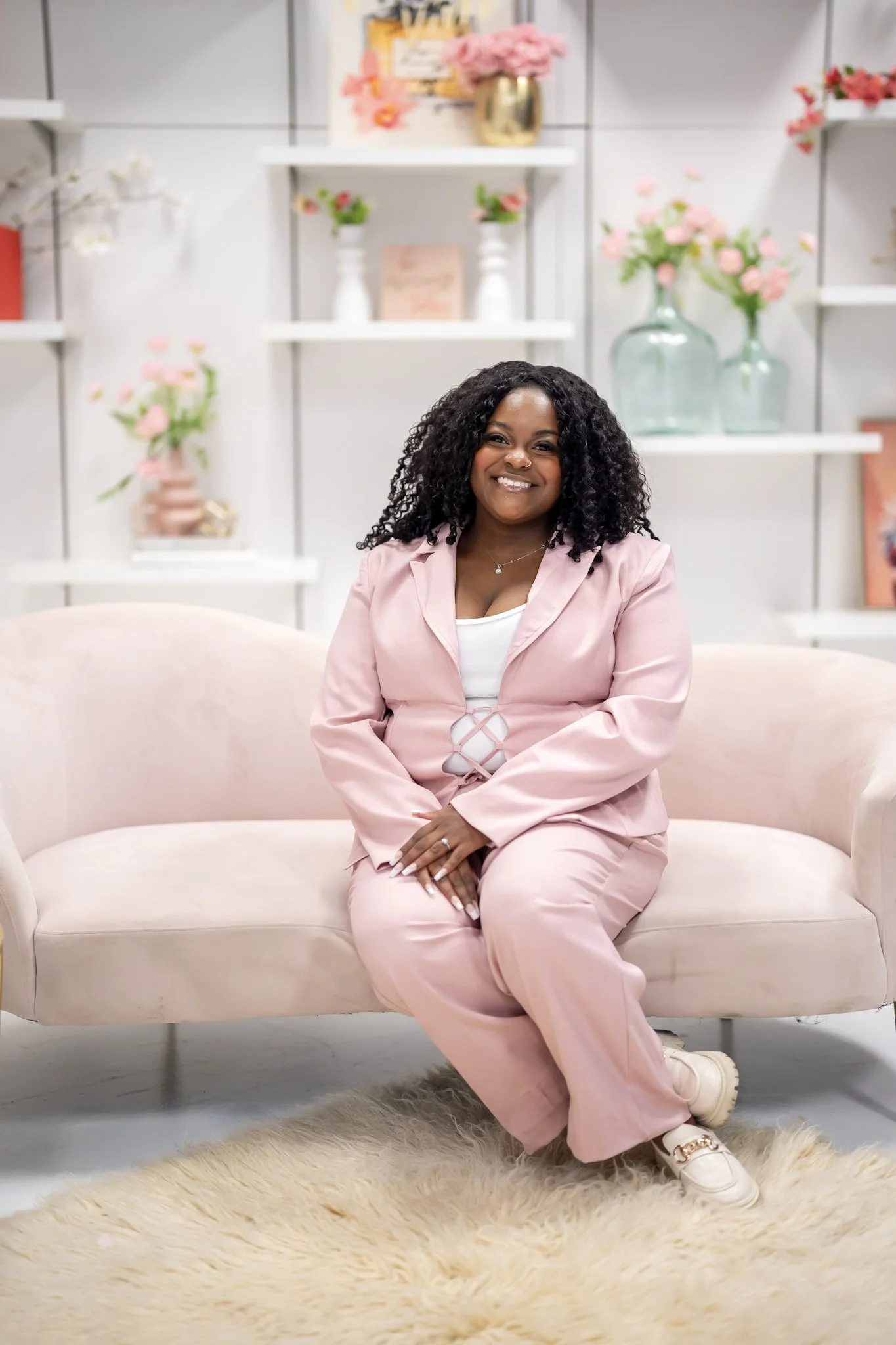 A woman with curly black hair sits on a light pink sofa in a room decorated with pink and white flowers, vases, and framed artwork.